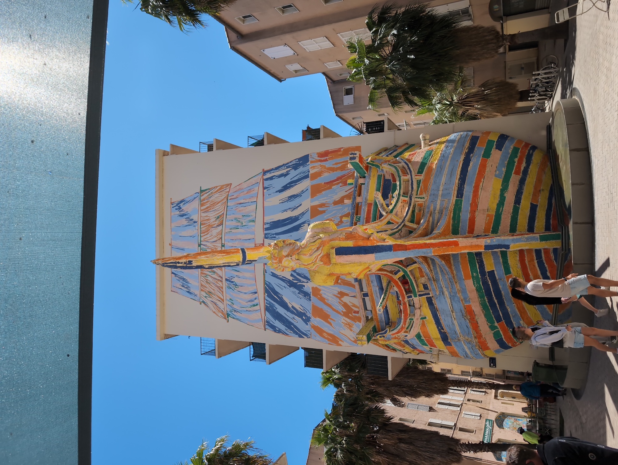 The image depicts a colorful mural on the side of a building in Toulon, France. The mural depicts a ship with sails and a figurehead, rendered in a vibrant, blocky style with horizontal stripes of blue, orange, yellow, and other colors. The sky is clear and blue.
Below the mural, on the street, are people. Two people are standing directly in front of the mural, looking up at it. Other people are walking by. Palm trees are visible along the street. Buildings can be seen on either side of the street. A glass-paneled structure is visible on the left side of the image, possibly part of a storefront or building entrance.