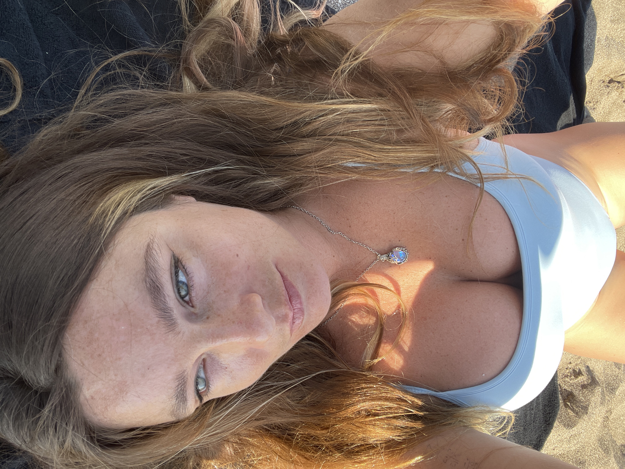 An upside-down, close-up, eye-level shot shows a fair-skinned woman lying on a dark towel on a sandy beach. Her long, wavy, auburn hair is spread around her face, which is tilted to her right. She is wearing a light blue bikini top, with the cleavage of her breasts visible. A silver necklace with a moonstone pendant rests between her breasts. The background is blurred sand and beach.