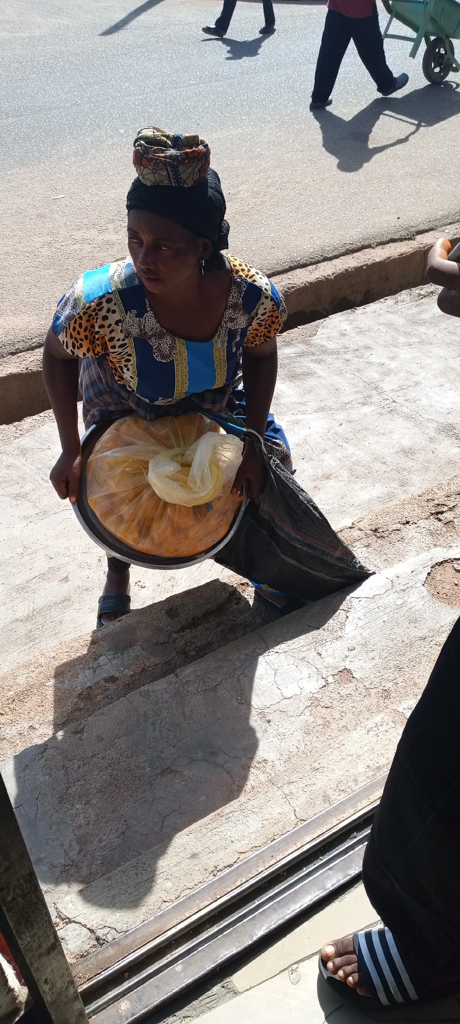 This outdoor image, taken in Jos, Nigeria, depicts a woman, likely a street vendor, in a moment of pause on a bright, sunny day. She is seen from an elevated perspective, crouching or resting on a concrete step or raised platform beside a street.
The woman wears a vibrant, short-sleeved top featuring blue, gold, and leopard print patterns, paired with a dark headwrap. Perched atop her headwrap is a smaller, decorative patterned cloth bundle, possibly a support pad for carrying items on her head. She holds a large, round metallic tray containing a substantial quantity of yellowish food items, protected within clear and yellow plastic bags. A dark, possibly woven bag is also visible, slung over her shoulder or resting near her. Her expression is neutral and focused.
The background shows an asphalt road bathed in sunlight, where strong shadows are cast by passersby. To the left, the legs and shadow of one person are visible. To the right, another person, wearing a red shirt and dark trousers, pushes a green wheelbarrow. In the immediate foreground, at the bottom right, a portion of a person's leg and foot, clad in a striped slide sandal, is visible.
The scene is set in an urban environment, characterized by concrete ground with visible cracks and a curb separating the sidewalk from the road. The bright light and distinct shadows indicate it is daytime. The image captures a snapshot of daily life and street commerce. No discernible text is visible.