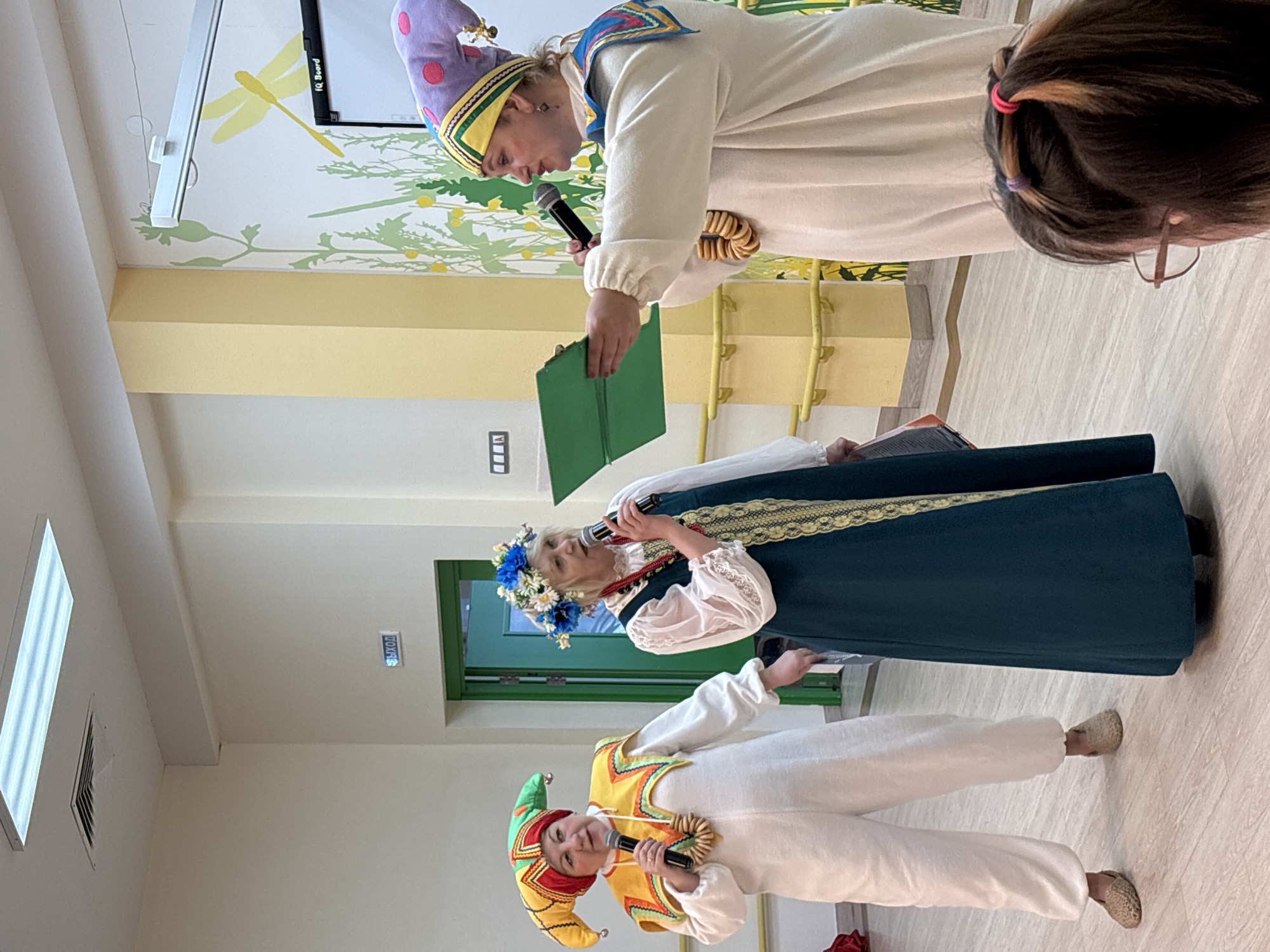 Three people are onstage indoors in Izhevsk, Russia. The person closest to the camera wears a yellow and green hat with hanging tassels and a white costume with a yellow and orange vest; they hold a microphone. The person in the middle wears a blue dress with a golden trim, white blouse, and a flower crown; they hold a microphone. The person in the background wears a white costume and a purple and yellow hat with a blue border; they hold a book and a microphone. A stack of bagels is on their shoulder. Behind them is a wall with a mural of green plants and a yellow dragonfly.