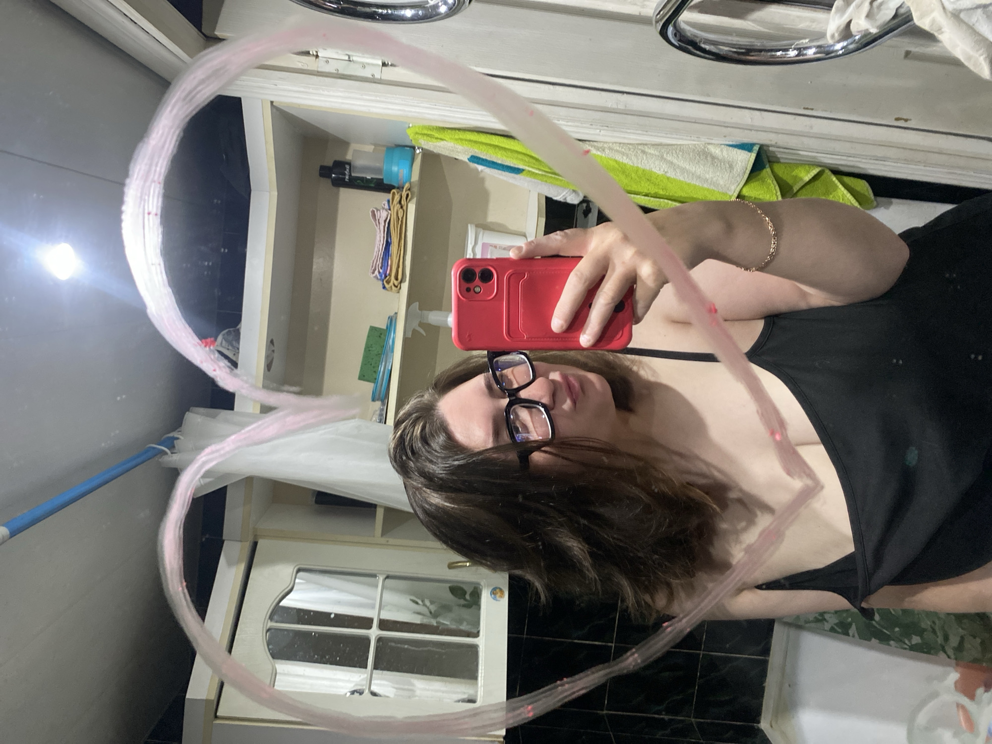 A mirror selfie taken in a bathroom. The reflection captures a person wearing glasses and a black tank top, holding a red smartphone. The mirror is decorated with a drawn pink heart. Background shows a bathroom setting with cabinets, a towel, and various toiletries. The location is Chaikovsky, Russia.