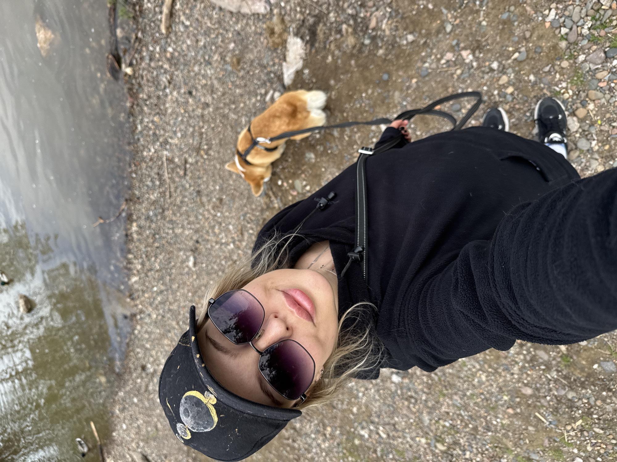 A person takes a selfie while walking a Corgi on a leash near a body of water. The person is wearing a black hoodie, sunglasses, and a black baseball cap with a gold moon design. The Corgi is orange and white. The ground is covered in gravel and pebbles. The water appears murky. The location is likely near a body of water in Krasnoyarsk, Russia.