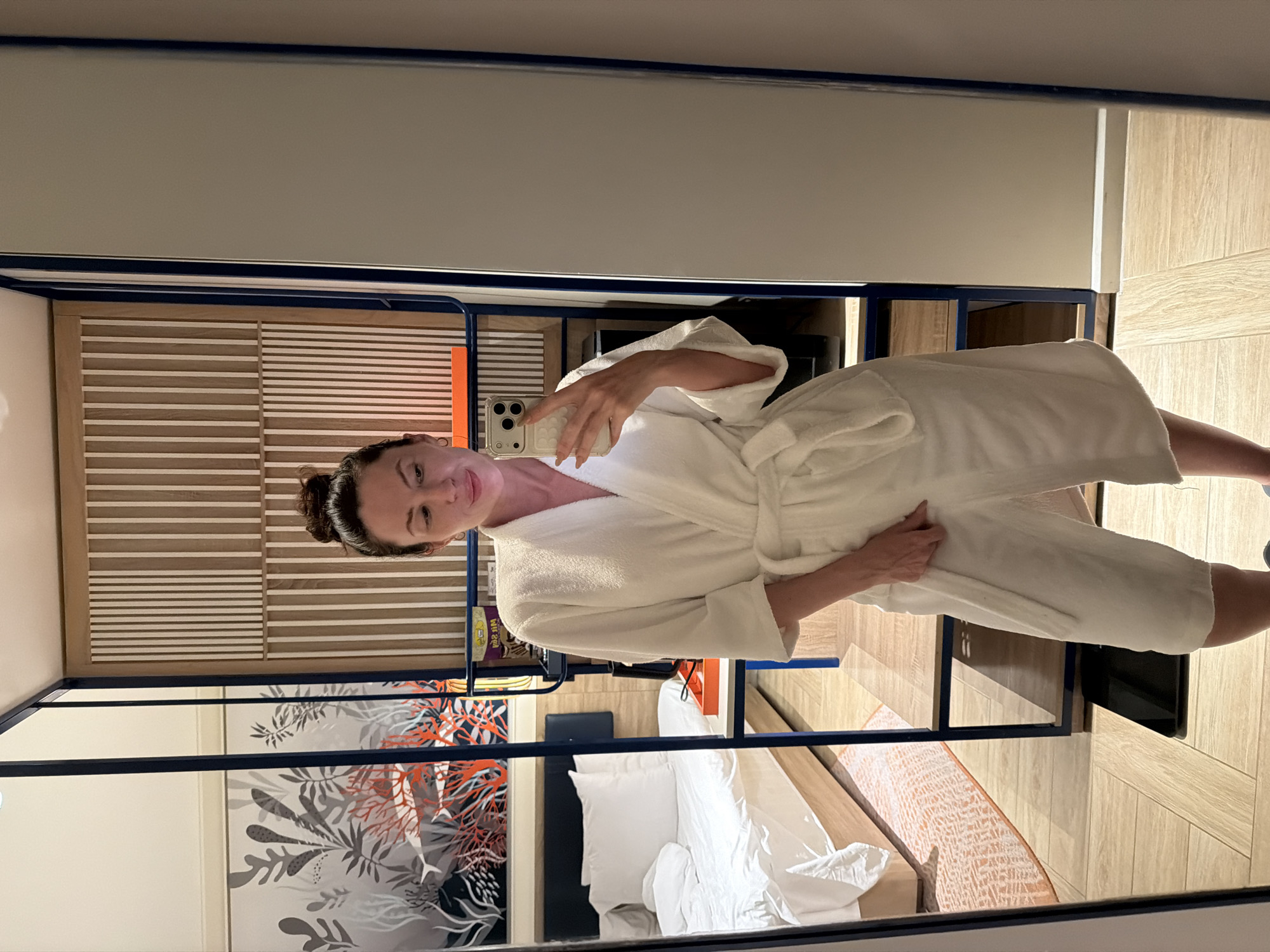 An adult female is depicted taking a mirror selfie in a room in Dong Ninh Hoa Ward, Vietnam. She is positioned centrally in the frame, wearing a white terrycloth bathrobe tied at the waist. Her dark hair is gathered into a top bun. She holds a light-colored smartphone with multiple rear cameras in her right hand, pointed towards the mirror. Her left hand rests on the front of the bathrobe. She exhibits a slight smile and a closed right eye. Small hoop earrings are visible on her ears.
The floor of the room features light-toned wooden planks arranged in a staggered pattern. A full-length mirror, framed in dark blue, captures the scene. To the left, reflected in the mirror, is a bed with white bedding, two white pillows, and a dark blue headboard. Above the bed, a wall mural displays stylized marine life, including red coral structures, gray and white plant-like forms, and dark outlines of fish against a light background. An orange tray is placed on a dark blue metal-framed table positioned next to the bed. A patterned rug, predominantly orange, is partially visible beneath the bed. To the woman's right, beyond the mirror's edge, a vertical slatted wooden panel with white strips is visible, along with parts of a dark blue metal shelving unit. A dark, flat rectangular object, possibly a weighing scale, rests on the floor near the bottom right.