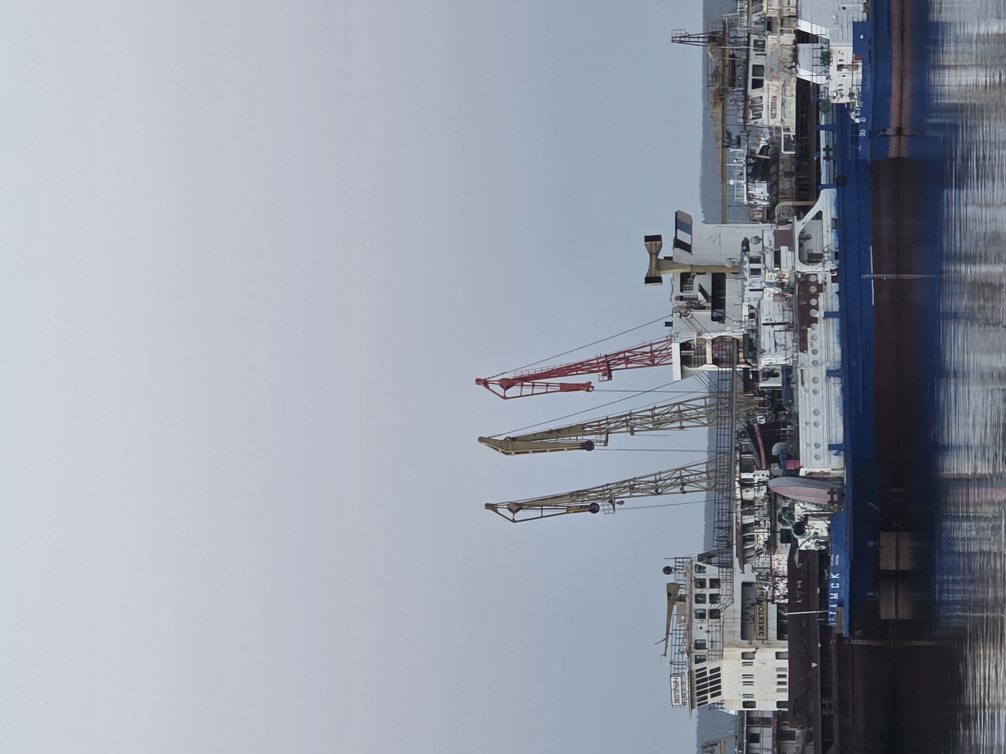 The image shows a ship docked near the city of Chaikovsky, Russia. The ship, painted in white, blue, and black, features three prominent cranes on its deck, one red and two yellow. The ship's name, "ЭЖЕКТОРЫ ПЕТАМСК" (Ejektory Petamsk), is visible. The background includes what appears to be a shipyard with other ships or industrial structures. The sky is overcast. The water reflects the ship and surrounding structures, indicating a calm or still body of water.