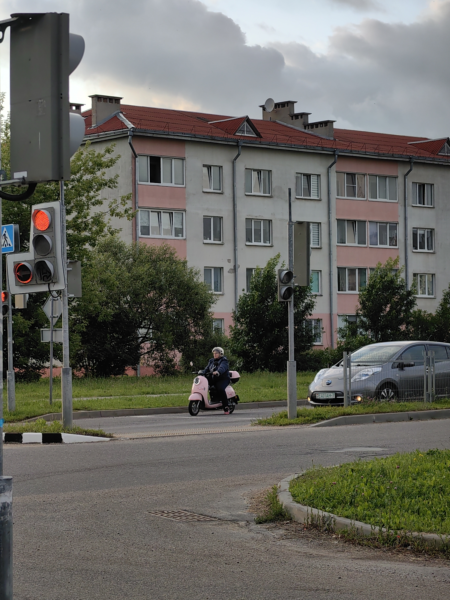 A person rides a pink scooter on a street in Fanipal, Belarus. The person is wearing a dark jacket and a white helmet. The scooter is moving towards the right of the image. In the background, there is a large apartment building with a red roof and pale pink and gray walls. A silver car is parked on the right side of the street, facing the viewer. Traffic lights are visible on the left side of the image, with the red light illuminated. The scene is set on a cloudy day, with lush green grass and trees lining the street. The road is asphalt.