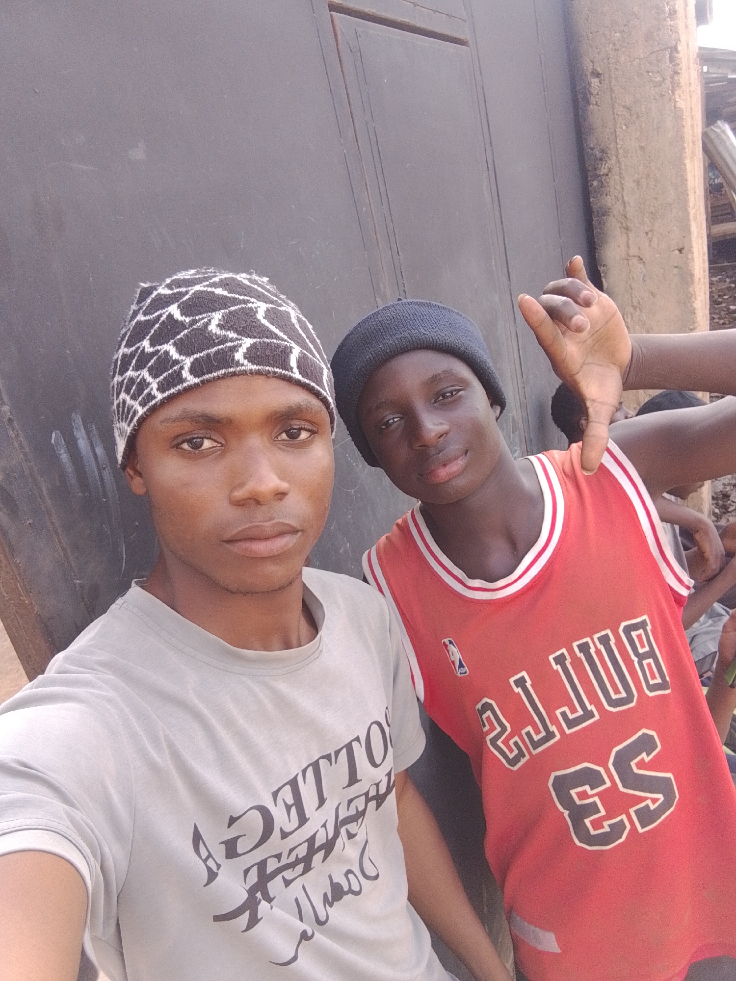 Two young men, likely of West African descent, are pictured outdoors in a dusty, urban environment. The man on the left, wearing a black and white patterned beanie and a light grey t-shirt, is looking directly at the camera with a neutral expression. He has dark eyes and skin. The man on the right, wearing a dark beanie and a red Chicago Bulls basketball jersey with the number "23" and "BULLS" visible, is looking slightly off-camera and making a hand gesture with his right hand. His skin tone is darker than the man on the left. Behind them, a dark, weathered metal door or wall serves as a backdrop. To the right of the door, a rough, concrete structure is visible, with glimpses of what appear to be makeshift structures or debris in the distance, suggesting a less developed part of the city. The overall scene implies a casual, candid moment captured on a street or in a backyard area. The image does not provide explicit visual cues to pinpoint Bukuru, Nigeria, beyond the general impression of a developing urban area.