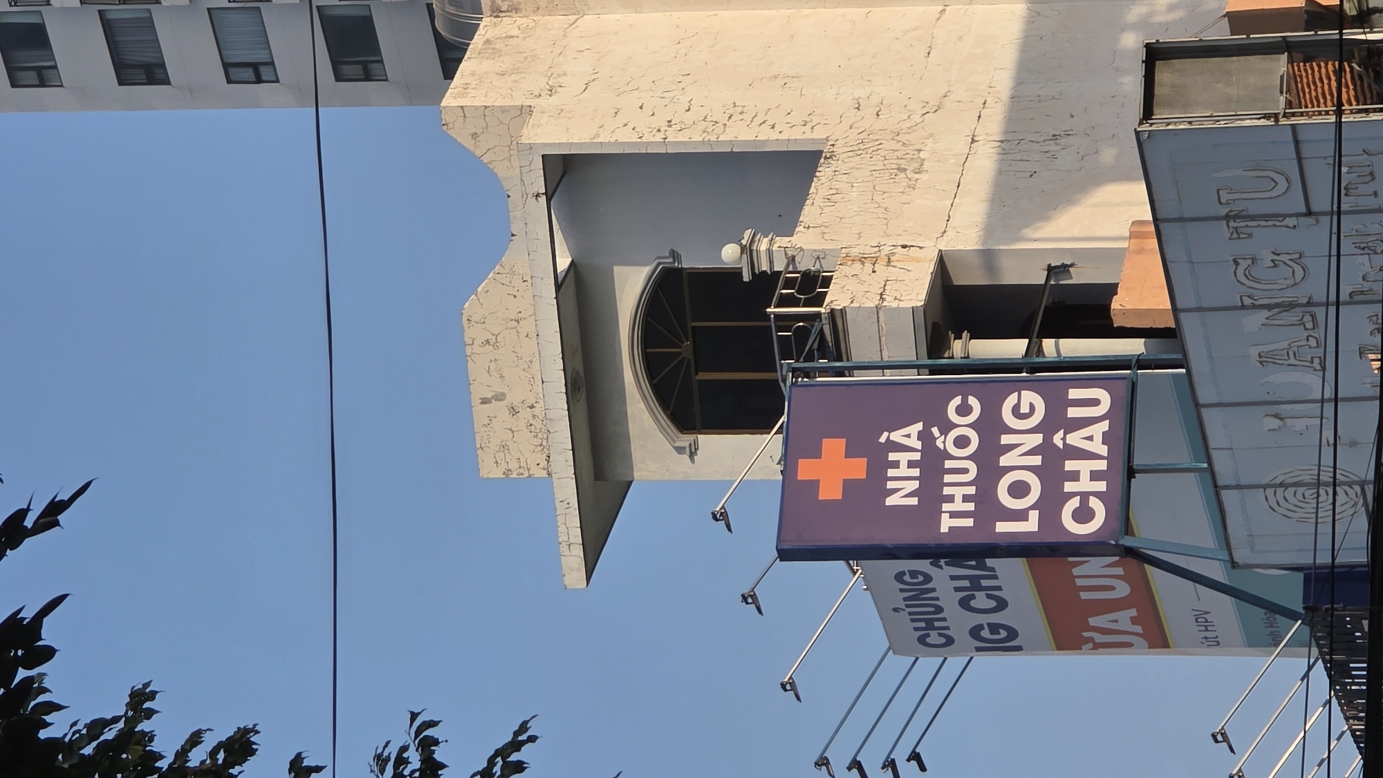 The image is a photograph taken from a low angle, capturing the exterior of a building in what appears to be Nha Trang, Vietnam. Dominating the frame is a blue sign with the text "NHÀ THUỐC LONG CHÂU" (Long Chau Pharmacy), accompanied by a red cross symbol. Below this, other signage elements are present with partially obscured text such as "CHÚNG IG CHÂ," "DA UN," and "út HPV ánh Hòa."
The building behind the signage features a unique architectural design with a semicircular window and textured, light-colored walls. In the distance, the top of another building with multiple windows can be seen. A power line stretches across the image, and some trees with dense foliage are visible in the lower part of the photo. The sky is a clear, bright blue, suggesting a sunny day.