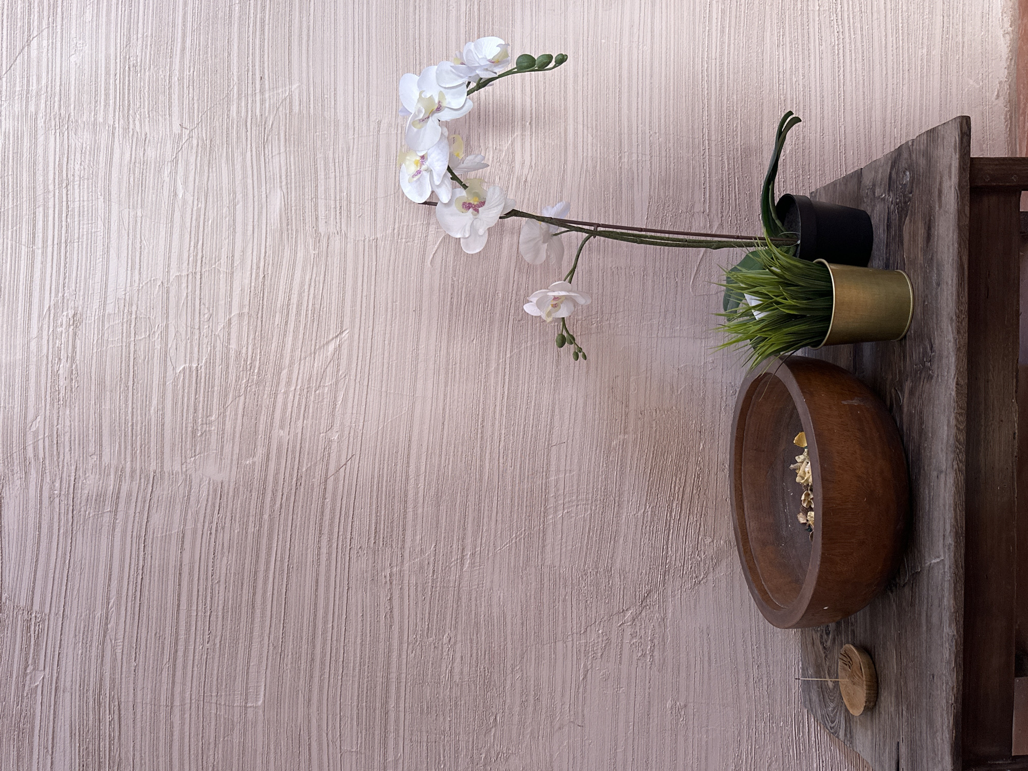 A horizontal indoor shot features a textured wall on the left, transitioning to a wooden shelf arrangement on the right. The wall is a light, creamy color with a vertical, striated texture. On the right side, a rustic wooden shelf displays a sparse collection of items. An arrangement of faux white orchids extends from the shelf onto the wall. Two small pots, one black and one brass-colored, contain artificial greenery. Below the pots, a large wooden bowl sits on the shelf, containing what appears to be dried petals or potpourri. A small, round wooden object is affixed to the shelf beneath the bowl.
While the prompt suggested the location is in North Kuta, Indonesia, there is no information in the provided image that confirms this.