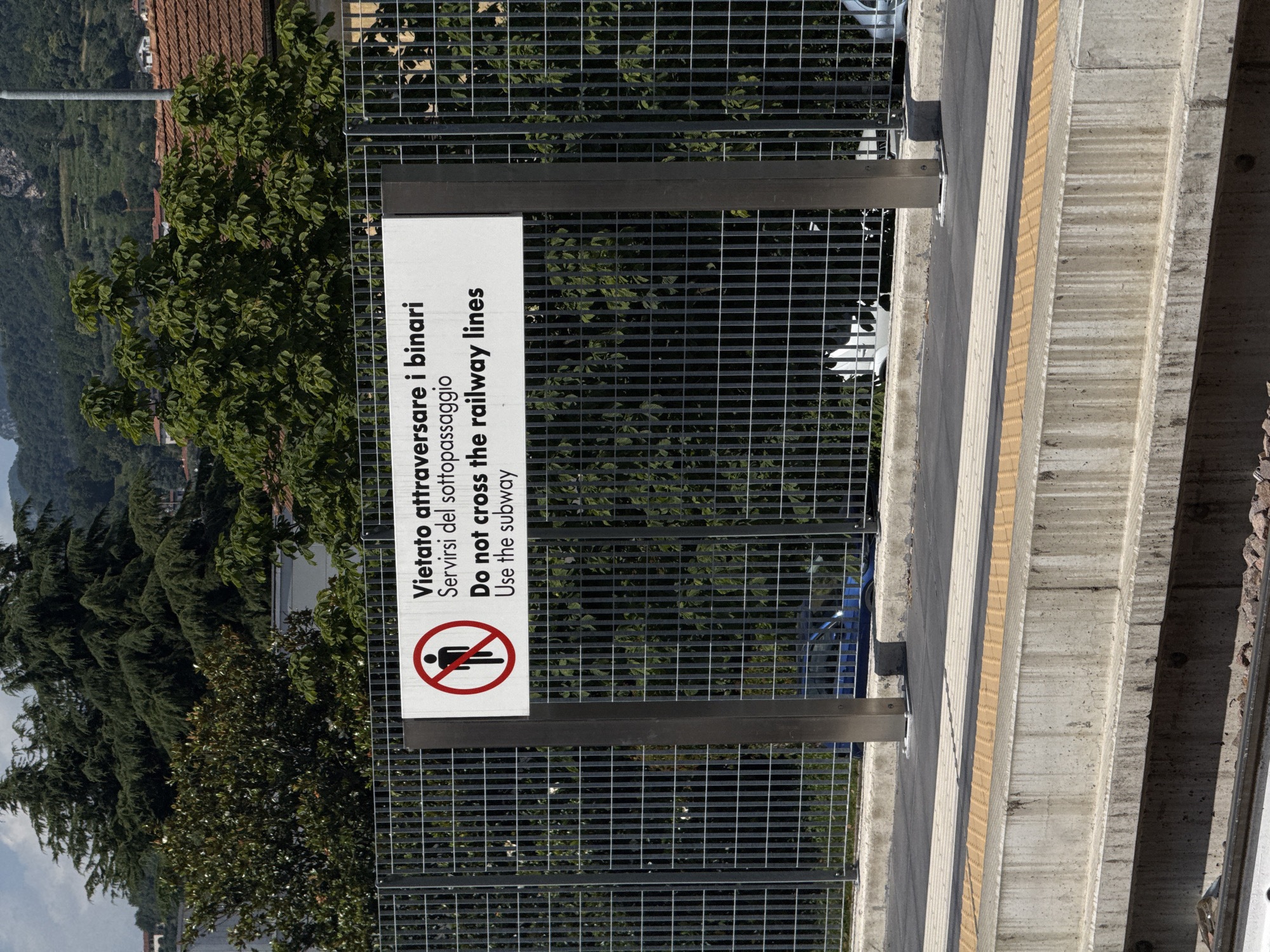 An eye-level, medium shot captures a sign in front of a metal fence at a train station. The sign, in Italian and English, reads "Vietato attraversare i binari Servirsi del sottopassaggio" and "Do not cross the railway lines Use the subway". To the left of the text is a red circle with a diagonal line through it, enclosing a black silhouette of a person. Behind the fence, trees and buildings are visible under a cloudy sky. The foreground shows the train platform with a yellow tactile paving strip along the edge, and a concrete structure below.