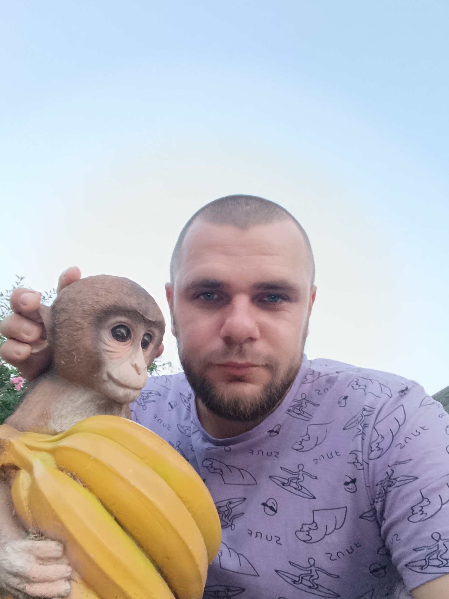 A close-up, low-angle selfie shows a Caucasian man with a shaved head and a beard. He is wearing a light purple t-shirt with a repeating pattern of surfers. The man is holding a statue of a monkey with bananas. The monkey statue is to the left of the man and is a light brown color. It has large, dark eyes and appears to be smiling. The bananas are a bright yellow and are positioned in front of the monkey. The background of the image is a bright, clear sky, suggesting an outdoor setting, possibly during daylight. There are some hints of green foliage and a pink flower on the left side of the image, indicating a garden or park environment. The image appears to be taken in Unknown, Unknown.
