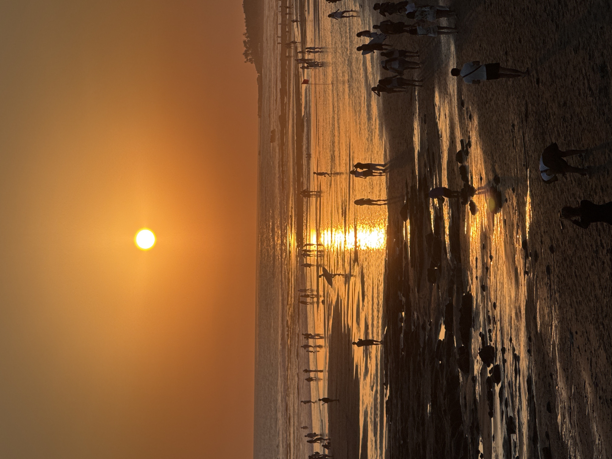 The image depicts a sunset at a beach in North Kuta, Indonesia. Silhouettes of numerous people are visible on the sandy shore and in the shallow water. The sun, a bright orb, hangs in the orange sky, casting a shimmering reflection on the ocean surface. Some individuals are walking, while others appear to be gathered in groups. A few are holding surfboards, suggesting recreational activities. The foreground shows dark, rocky patches interspersed with wet sand, reflecting the warm hues of the sunset. The overall scene conveys a peaceful and lively atmosphere as the day comes to a close.