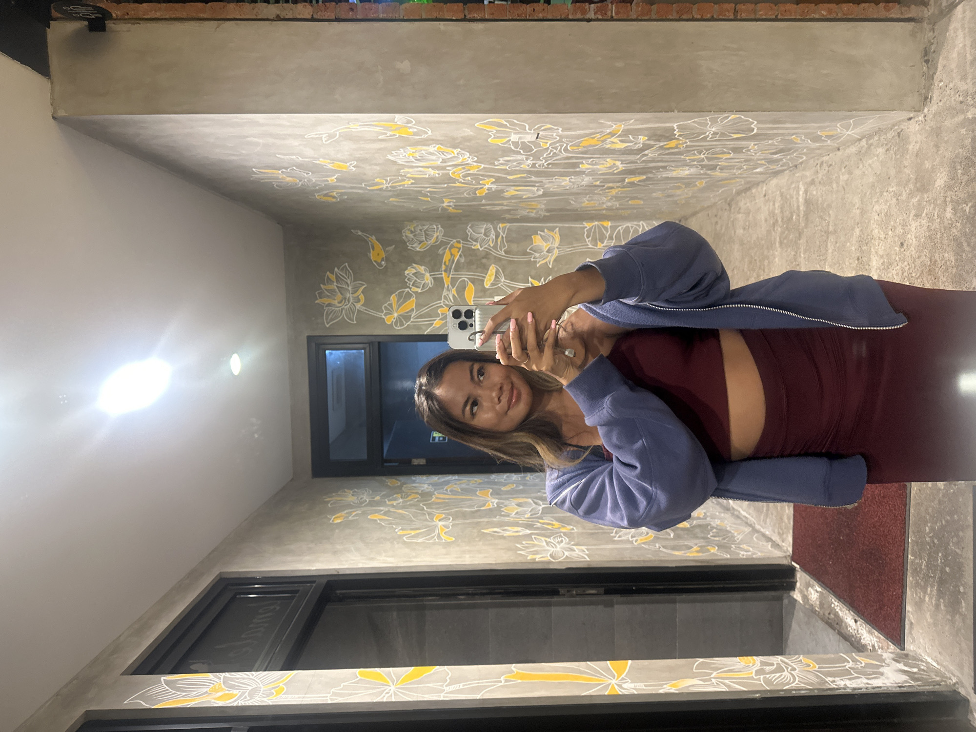 A young woman, appearing to be of Southeast Asian descent, stands in a narrow hallway and takes a mirror selfie with her smartphone. She is wearing a dark red crop top and matching ruched skirt, with an open blue hooded jacket layered over the top. Her long, brown hair is down, and she is looking at her phone. The hallway has concrete walls decorated with a repeated pattern of white and yellow lotus flowers and fish. To the left of the woman is a dark doorway with what appears to be a reflection of a tiled surface inside. A dark red mat is on the floor in front of the doorway. To the right of the woman, the hallway continues, with a brick wall visible at the far end. The lighting in the hallway is bright, likely from an overhead light source. Based on the wall art and architectural style, this location is likely a boutique hotel or guesthouse in the city of Denpasar, Indonesia, possibly in a district known for its artistic or spiritual ambiance, such as near the Pura Besakih or in a more bohemian area.