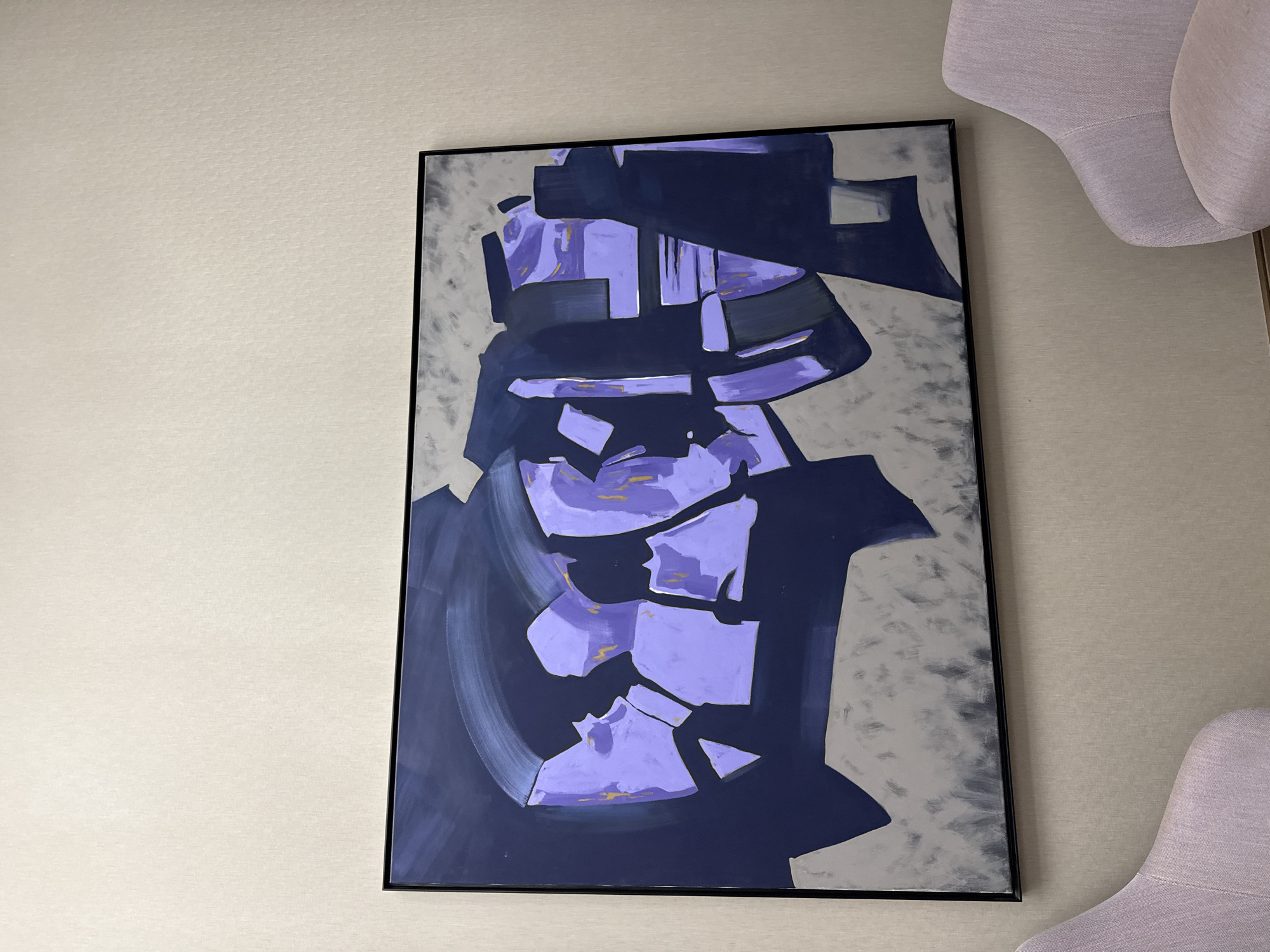 A vertical shot captures a contemporary indoor setting, dominated by a large abstract painting on a light-colored wall. The painting, encased in a thin black frame, features a dynamic composition of fragmented geometric and organic shapes. Shades of deep navy blue, vibrant purple, and muted gray define the artwork, with the purple sections often appearing as broken blocks accented by subtle hints of gold or yellow. The background of the painting is a textured gray, giving a painterly depth.
To the right of the painting, partial views of two upholstered chairs or armchairs are visible. They are a soft lilac or lavender color, harmonizing with the purple tones in the artwork. The wall itself has a subtle, woven or textured pattern in an off-white or light beige color.
No specific activity is taking place; the image is a still depiction of interior decor. There are no people visible, nor any discernible text. The time of day and external weather conditions cannot be determined from the indoor lighting. The scene suggests a modern and stylish interior, with the context provided indicating its location in Khimki, Russia.