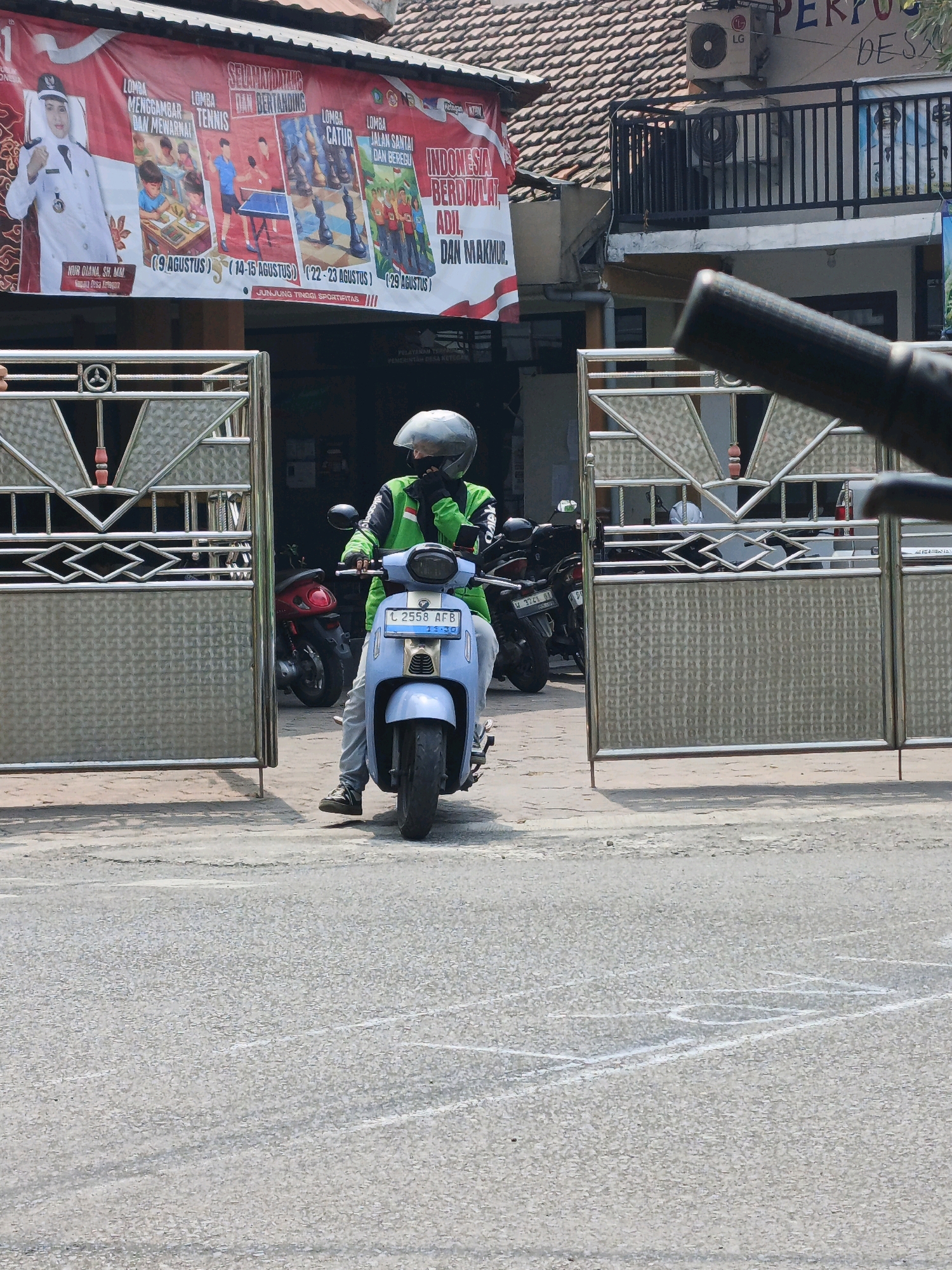The image captures a street scene in Ketegan, Indonesia, with a primary focus on a person on a light blue scooter. The rider is wearing a green jacket, black gloves, and a silver helmet, with their face obscured by the helmet and a dark face covering. They are positioned in front of a large, ornate metal gate, which is partially open, revealing a dark interior with several parked motorcycles. To the left of the gate, a red banner with text and images is prominently displayed. The banner features a portrait of a woman in a white uniform, along with illustrations of children drawing, table tennis, chess pieces, and a group of children running. Text on the banner is in Indonesian and mentions events like "Lomba Menggambar dan Mewarnai" (Drawing and Coloring Competition), "Lomba Tenis" (Tennis Competition), "Lomba Catur" (Chess Competition), and "Lomba Jalan Santai dan Berlari" (Leisure Walk and Running Competition). The scooter has a license plate that reads "L 2558 AFB". In the background, a building with a tiled roof and an air conditioning unit is visible. The ground in front of the gate is asphalt, with some white chalk markings that appear to form a game or a diagram. The overall scene suggests a public space, possibly near a community center or a government building, given the banner's content and the woman's depicted uniform.