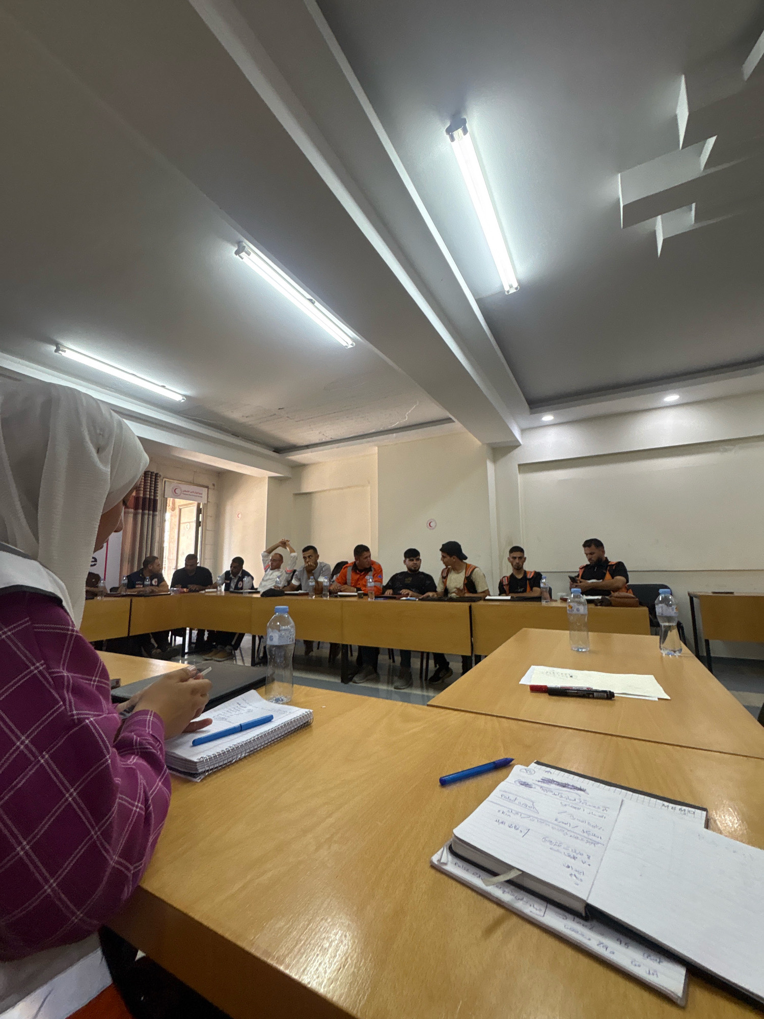 In a brightly lit room, likely a classroom or meeting hall, a group of people are seated around a large U-shaped table, engaged in what appears to be a training session or discussion. The room has a dropped ceiling with fluorescent lights. On the table in the foreground, a notebook with handwritten notes and a blue pen are visible. Behind it, another notebook and a red marker lie on a piece of paper. A water bottle is also present on the table.
The individuals around the table are diverse in age and attire. Some are wearing casual clothing, while others are in work uniforms, including vests. One person in the foreground, wearing a headscarf and a plaid shirt, is interacting with the notebook. In the background, a red crescent emblem is visible on the wall, suggesting a connection to a humanitarian or health organization. The overall atmosphere seems to be one of focused activity. The presence of the red crescent emblem suggests the location could be within Gaza, Palestinian Territories.