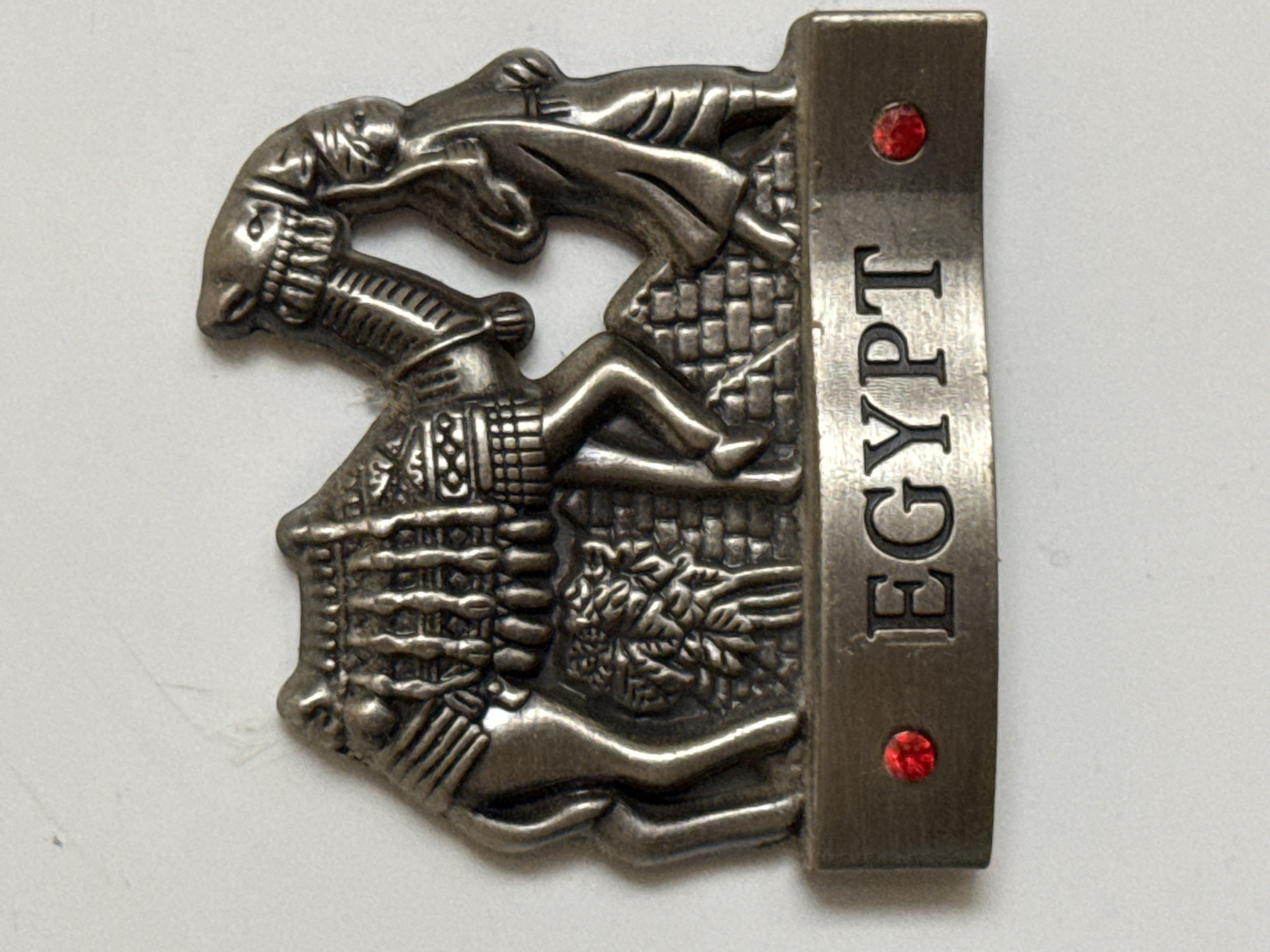 This is a close-up, top-down studio shot of a silver-colored metal souvenir item, likely a magnet or a pin, featuring an Egyptian motif. The item is placed on a plain white, slightly textured background.
The main subject of the souvenir is a stylized depiction of ancient Egyptian figures and symbols. On the left side, there is a figure of a pharaoh wearing a nemes headdress and an elaborate collar. Next to the pharaoh, a seated figure, possibly a queen, is visible. Below them, a scene unfolds with what appears to be a sarcophagus or a tomb structure, decorated with hieroglyphic-like patterns. A stylized depiction of lotus flowers or reeds is also incorporated into the design. The right side of the souvenir is a rectangular metal bar with the word "EGYPT" inscribed vertically in bold, uppercase letters. Two small, round, red gemstones are embedded in the metal bar, one above the text and one below.
The metal has a tarnished, antique silver appearance, highlighting the embossed details. The overall impression is of a decorative item celebrating Egyptian culture and history. There is no indication of time of day, weather, or specific emotions conveyed by the figures, as they are depicted in a static, symbolic manner. The only explicit textual information is the word "EGYPT" on the metallic strip.