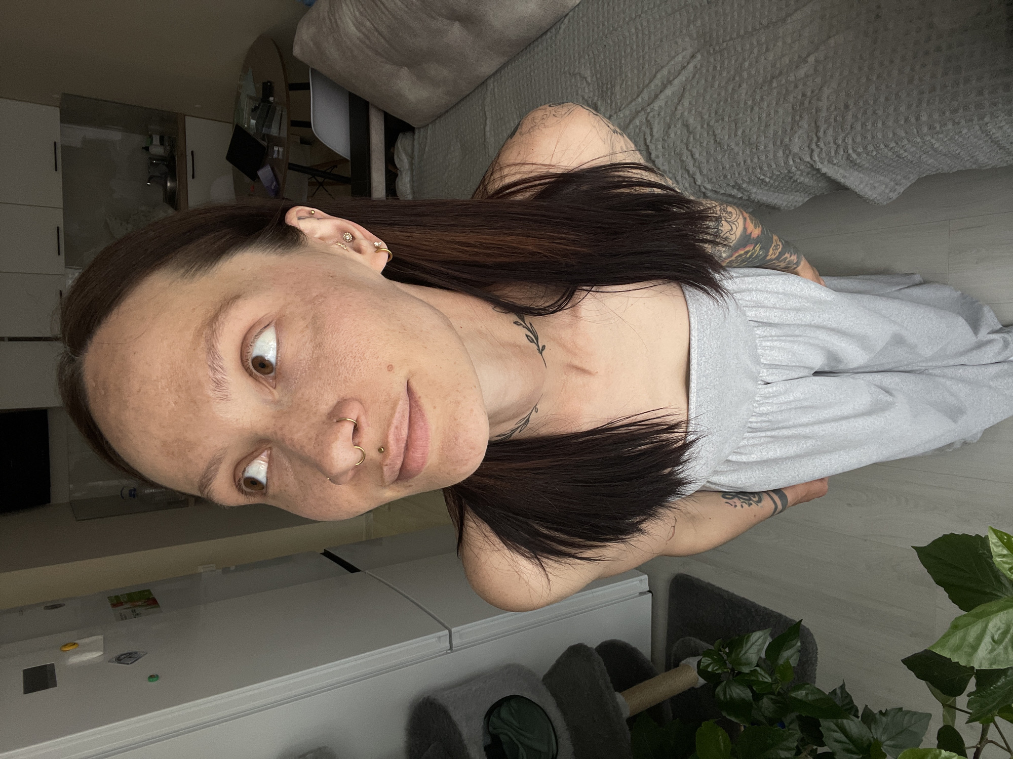 A woman is shown in a medium shot, tilted to the left. Her hair is long and dark brown, with a lighter brown tint and is parted in the middle. She is wearing a light grey dress. The woman has fair skin with freckles, brown eyes, and multiple piercings in her ear and nose. She has tattoos on her arms and neck.
The background shows part of an apartment or house interior. To the lower left is a white refrigerator, with magnets. Next to the refrigerator is a grey pet tree. In the lower right is a potted green plant. At the top is a grey couch, with grey linen. The floor is light grey wood.
The image does not confirm or deny a location in the city of Pervomaysky, Russia.
