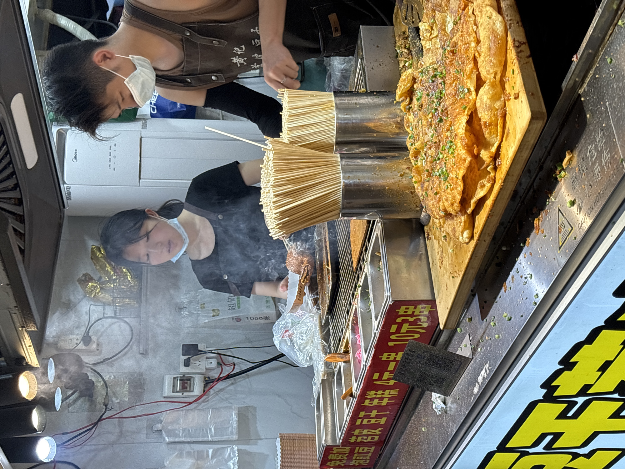 This is a medium shot, taken at an eye-level angle, of an outdoor food stall in Jiangjin, China. The stall is busy, with two vendors, a young woman and a young man, preparing food. The woman, wearing a light blue face mask, is on the left, holding a paper tray with food. She is standing behind a grill with skewers of food cooking on it, and steam is rising from the grill. The man, wearing a similar face mask and a brown apron, is on the right, serving food. He is also behind the grill, and appears to be handing a skewer to a customer.
In the foreground, there is a long metal tray with various toppings in compartments, and to the right of that, there is a wooden board with several flat, crispy food items, likely crepes or pancakes, sprinkled with green onions and spices. Behind the vendors, there is a wall with electrical outlets and wiring, and a white appliance that looks like a refrigerator or food warmer. There are also metal containers filled with wooden skewers.
The overall environment suggests a bustling night market or street food vendor area. The lighting is somewhat dim, with spotlights illuminating the food preparation area. There is a sign in Chinese with yellow lettering on a blue background at the bottom of the image. The food stall appears to be set up to serve customers quickly and efficiently.