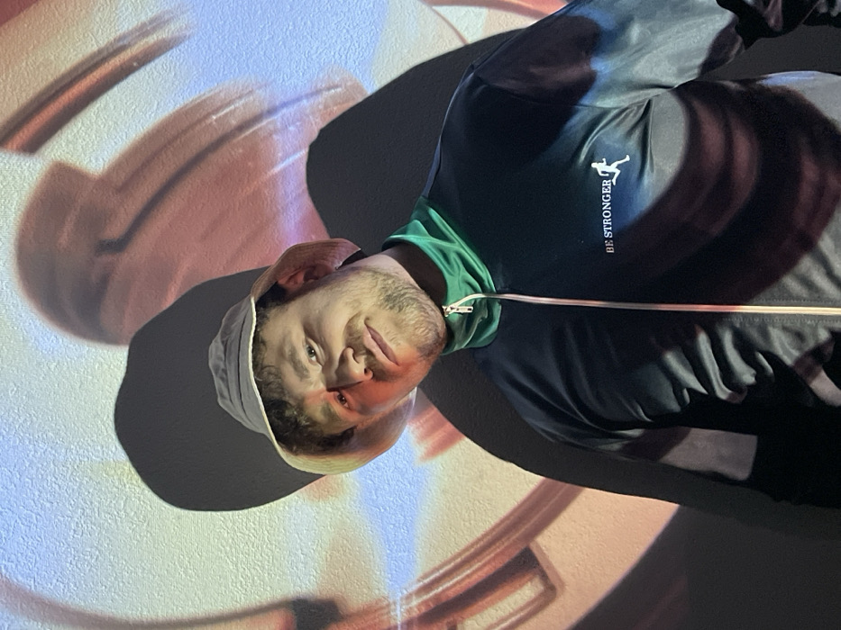 A man wears a white bucket hat, black jacket with green trim, and "BE STRONGER" logo. He stands against a wall illuminated with broad strokes of peach, white, and blue light. A large shadow is cast beside him. The image is rotated.