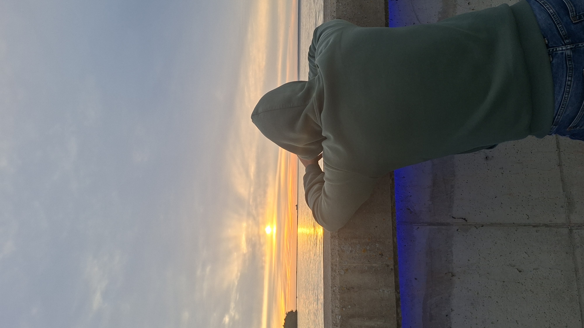A person wearing a dark green hooded sweatshirt and blue jeans leans over a concrete barrier, looking out at the sunset over the Baltic Sea. The sun, a bright orange orb, is low on the horizon, casting a golden glow across the water and sky. Wispy clouds streak across the sky, tinged with hues of orange, yellow, and pink. A distant ship is visible on the horizon. The concrete barrier has a strip of blue lighting along its edge. The scene appears to be a waterfront promenade or pier, likely in Tallinn, Estonia, given the clear view of the Baltic Sea at sunset.