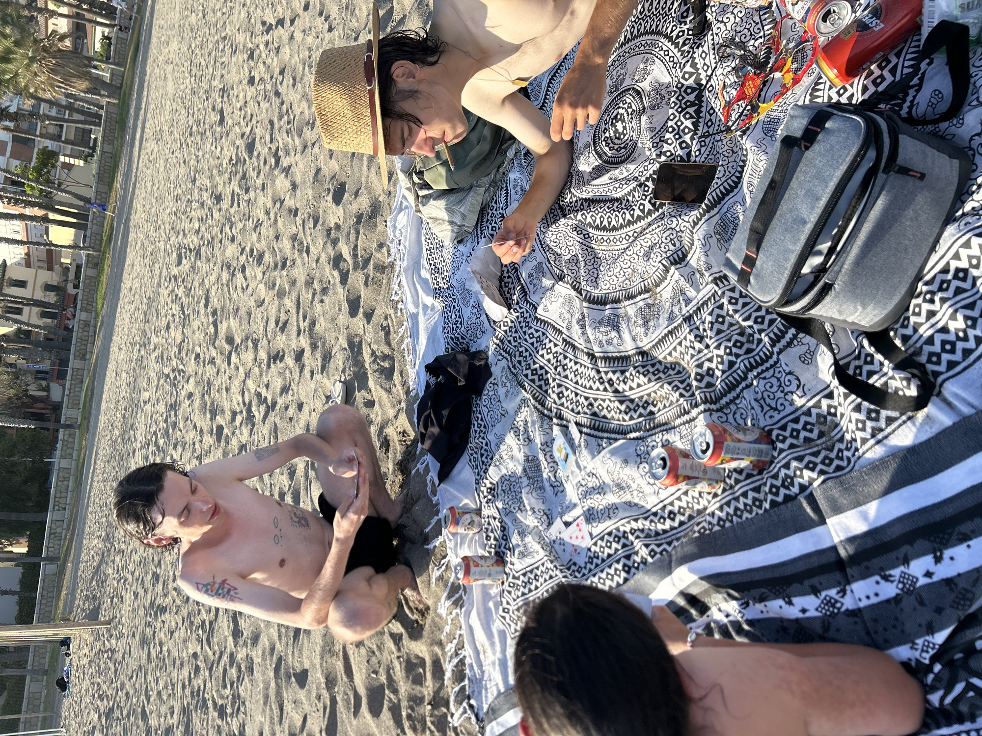 The image depicts three individuals gathered on a sandy beach, consistent with the coastline of Málaga, Spain, given the background architecture and vegetation. The scene appears to be set in the late afternoon or early evening due to the warm, low-angle sunlight.
In the center-top, a young male with dark hair, shirtless and wearing dark shorts, is crouched directly on the sand. He has multiple tattoos visible on his arms and chest. He is focused on a smartphone held in both hands. To the right, another young male, also shirtless, is lying prone on a black and white patterned blanket. He wears a straw fedora-style hat with a dark band and a gold-colored pendant around his neck. A thin, rolled item, resembling a cigarette, is held in his mouth, and he is holding a small, light-colored rectangular object, possibly a playing card, in his right hand. The back of a third person's head and upper shoulder, with dark hair, is visible in the bottom-left corner, indicating they are seated or kneeling on the blanket, facing away from the camera.
The patterned blanket, adorned with intricate mandala and animal motifs, serves as a central gathering point. Several aluminum beverage cans, some upright and some overturned, are scattered across the blanket. One yellow can clearly shows "Cerveza más fina," indicative of Coronita/Corona beer, and a red can displays part of a brand name, "ESPE," potentially Estrella Galicia. Playing cards, including a 5 of hearts and a 9 of spades, are spread on the blanket. A dark-colored smartphone lies face down near the bottom center, and a grey soft-sided cooler bag with black straps is positioned to the right. Colorful snack wrappers are also visible near the cooler bag.
The background features an expansive sandy beach with faint foot impressions. Beyond the sand, a low wall separates the beach from a paved promenade or walkway. A row of tall palm trees lines the promenade, behind which are multi-story buildings with balconies and varying architectural styles, characteristic of coastal urban areas in Málaga. A wooden post, likely part of a beach recreational setup, is visible on the far left of the beach. A white flip-flop lies in the sand near the first individual.