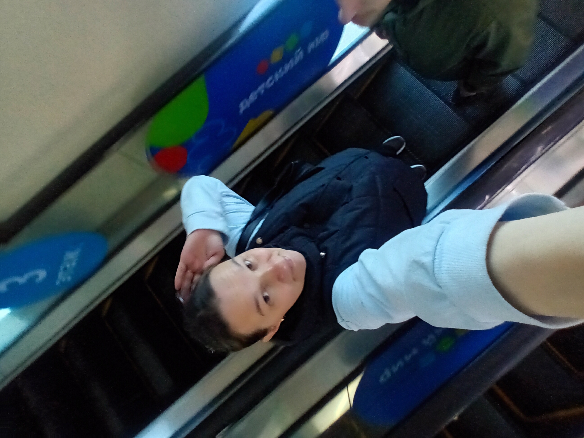 A person is taking a selfie while riding an escalator. The person is wearing a light blue long-sleeved shirt and a dark vest or jacket. The escalator has advertisements on the sides, including one for "Детский мир" (Detsky Mir), which translates to "Children's World." Another advertisement displays the number "3." The image suggests the person is in a shopping mall or public transit area. The location is likely Chaikovsky, Russia, based on the Russian text in the advertisements.