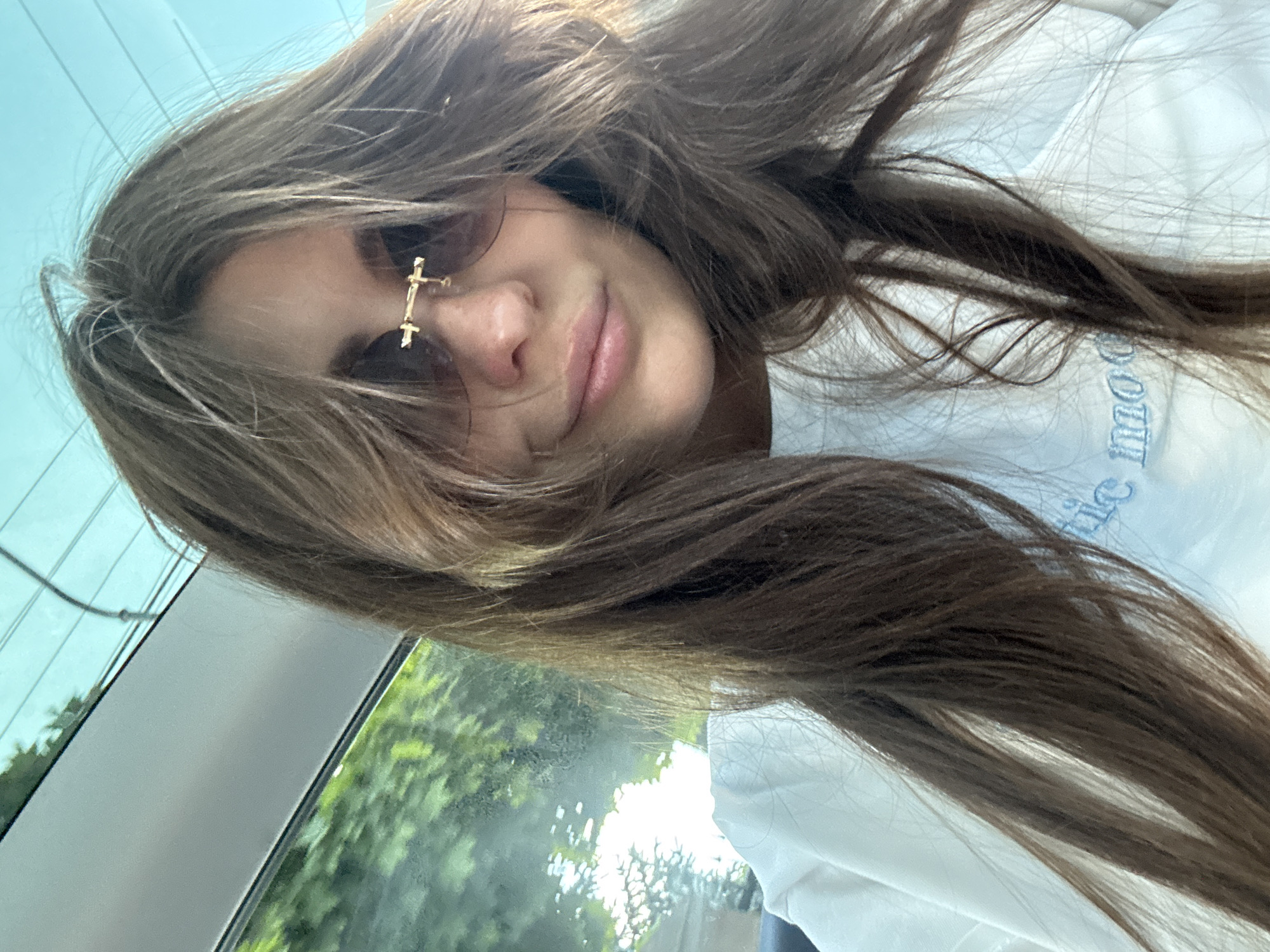 This is a close-up selfie of a young woman taken inside what appears to be a car in Sofiivska Borshchahivka, Ukraine, during the daytime. The woman is positioned with her head tilted, looking down towards the camera with a soft, relaxed smile. Her long, light brown hair, parted on the side, cascades around her face and shoulders, partially obscuring her right eye. She is wearing stylish round sunglasses with a distinctive gold cross-shaped detail on the bridge. Her left eye is visible, and she has natural makeup. She is dressed in a white t-shirt, which has light blue text partially visible on the right side of her chest, appearing to spell out "...mood" in a stylized font.
The background visible through the car window on the left side shows bright green foliage, indicative of trees or bushes, and a bright, possibly clear, sky above, suggesting sunny weather. Parts of the car's interior, such as the roof lining and window frame, are also visible. The lighting is bright, illuminating her face and hair. The overall mood of the image is casual and pleasant.