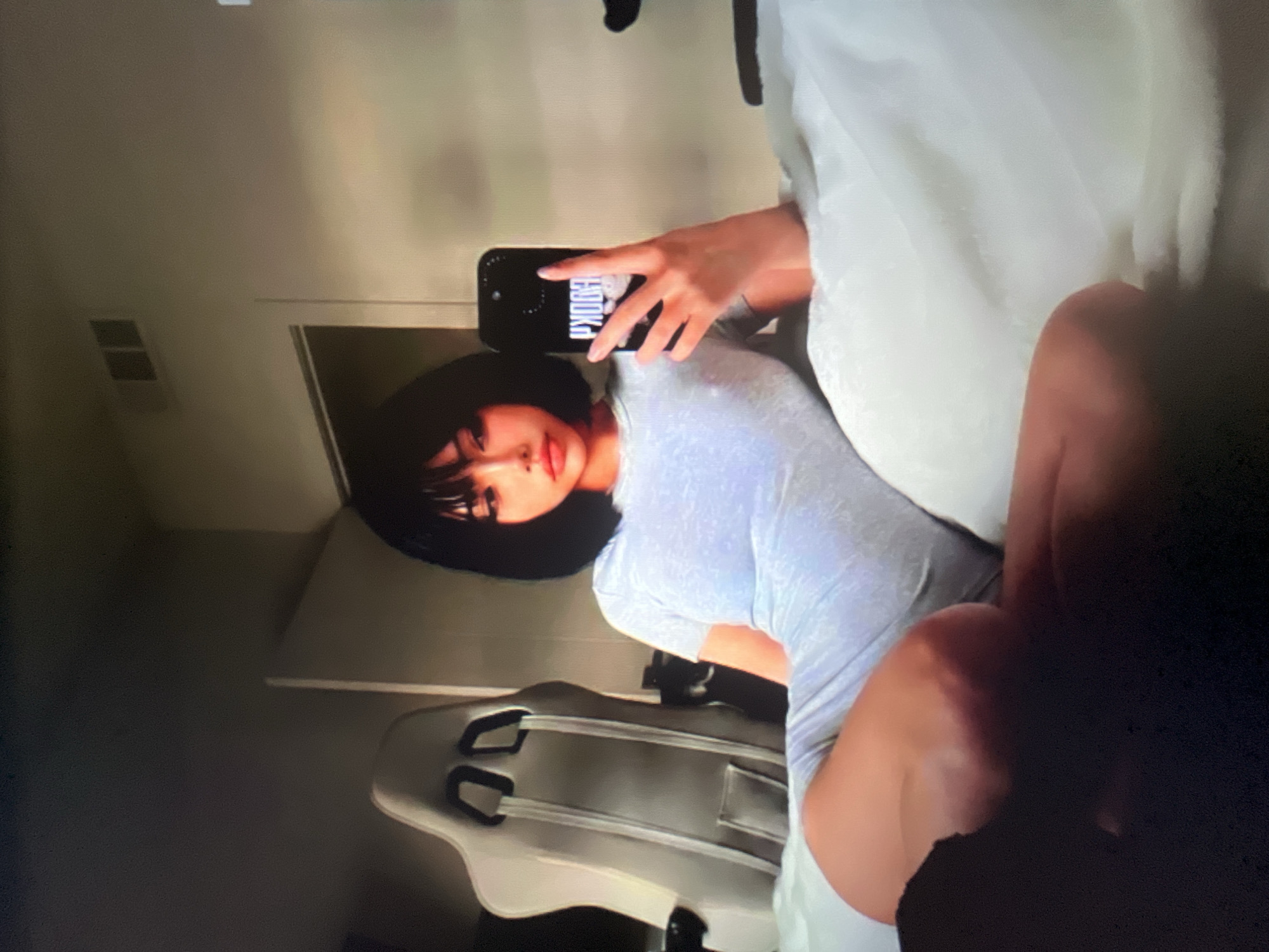 A young woman with short black hair and bangs, wearing a light blue short-sleeved romper, is lying on a bed and taking a selfie. She is holding a black smartphone in her right hand, and the phone case displays the word "BOOKA" in white lettering. Her left hand is resting on her thigh, which is partially obscured by a dark shadow. Behind her, to the left, is a white gaming chair with black accents. The room appears to be a bedroom, with a vent visible on the wall above the chair. The lighting is dim, with the woman's face and the phone being the most illuminated parts of the image.