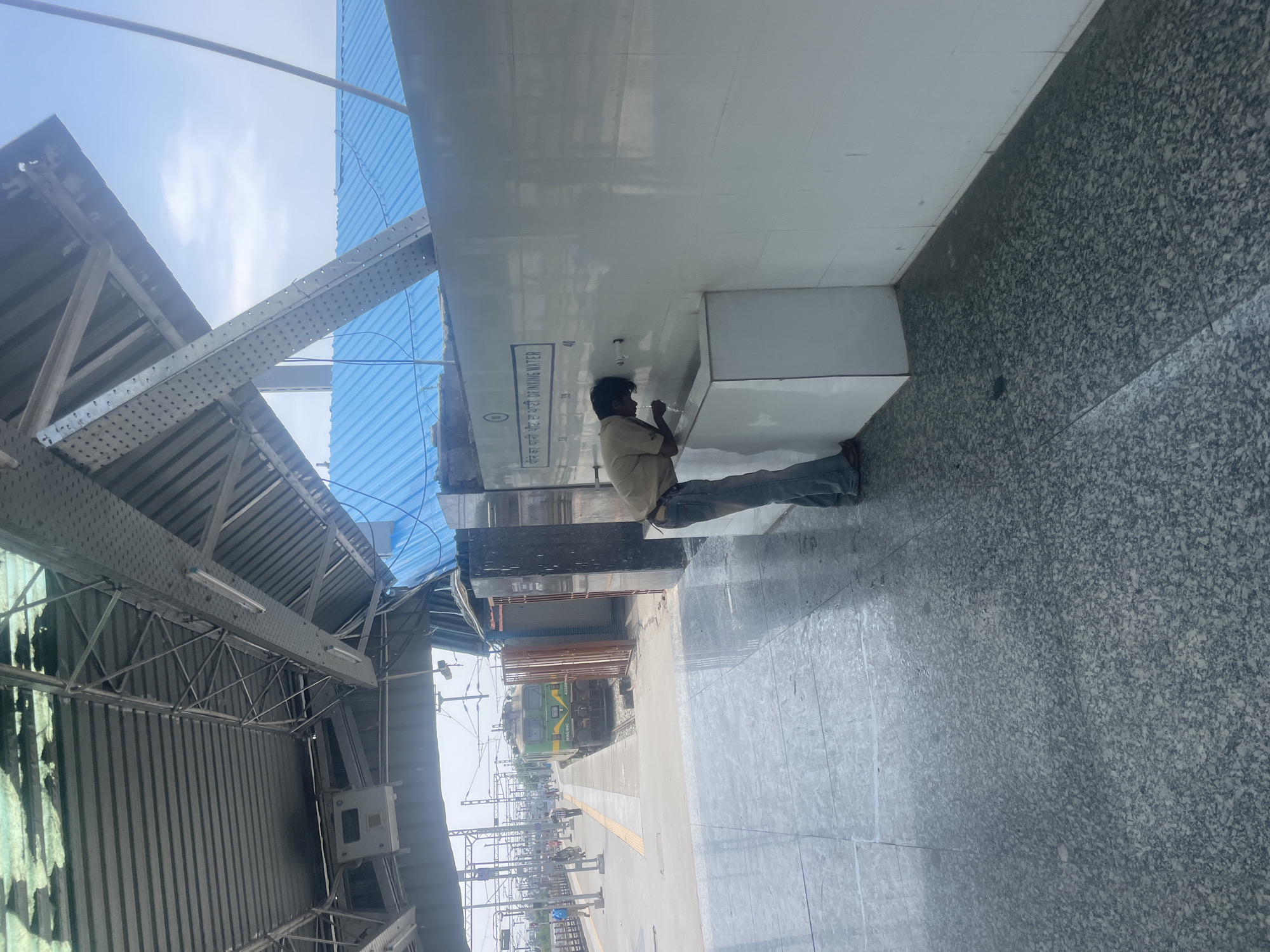 The image depicts a scene at the Pathankot Railway Station in Pathankot, India. In the foreground, a man wearing a light-colored shirt and blue jeans is drinking water from a public drinking fountain. The fountain is mounted on a white tiled wall, and the man is bending down to access the water. Behind him, a white basin is integrated into the wall. The floor is tiled with large, dark grey granite-like tiles, some of which appear wet.
In the middle ground, a green and yellow train is stopped at a platform. The train has the number "WAG-10H" visible on its side. Several people are walking on the platform in the distance. The platform has yellow tactile paving.
The background shows the overhead structure of the railway station, including a corrugated metal roof supported by metal beams. Beyond the station structure, a bright blue roof is visible, and the sky is light blue with some clouds. The overall environment suggests a sunny day at a busy train station.