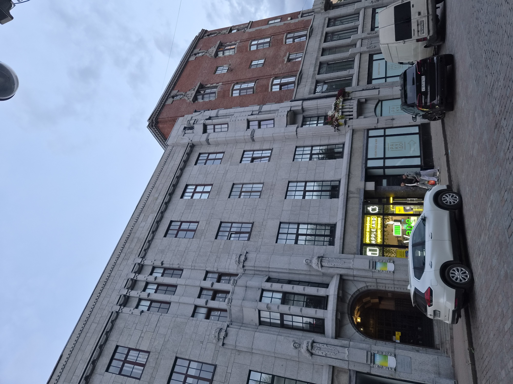 The image shows a street scene in Vyborg, Russia, featuring an old building with a granite facade and a brick addition. In front of the building, several cars are parked on a cobblestone street, including a white Nissan SUV, a black BMW SUV, and a white van. The building has large windows and decorative architectural elements, including statues on the granite section. A storefront with neon signs that read "APPLE AND" is visible, suggesting a retail establishment. Two people are walking on the sidewalk in front of another storefront labeled "ASIAN FOOD & BAR". The sky is cloudy.