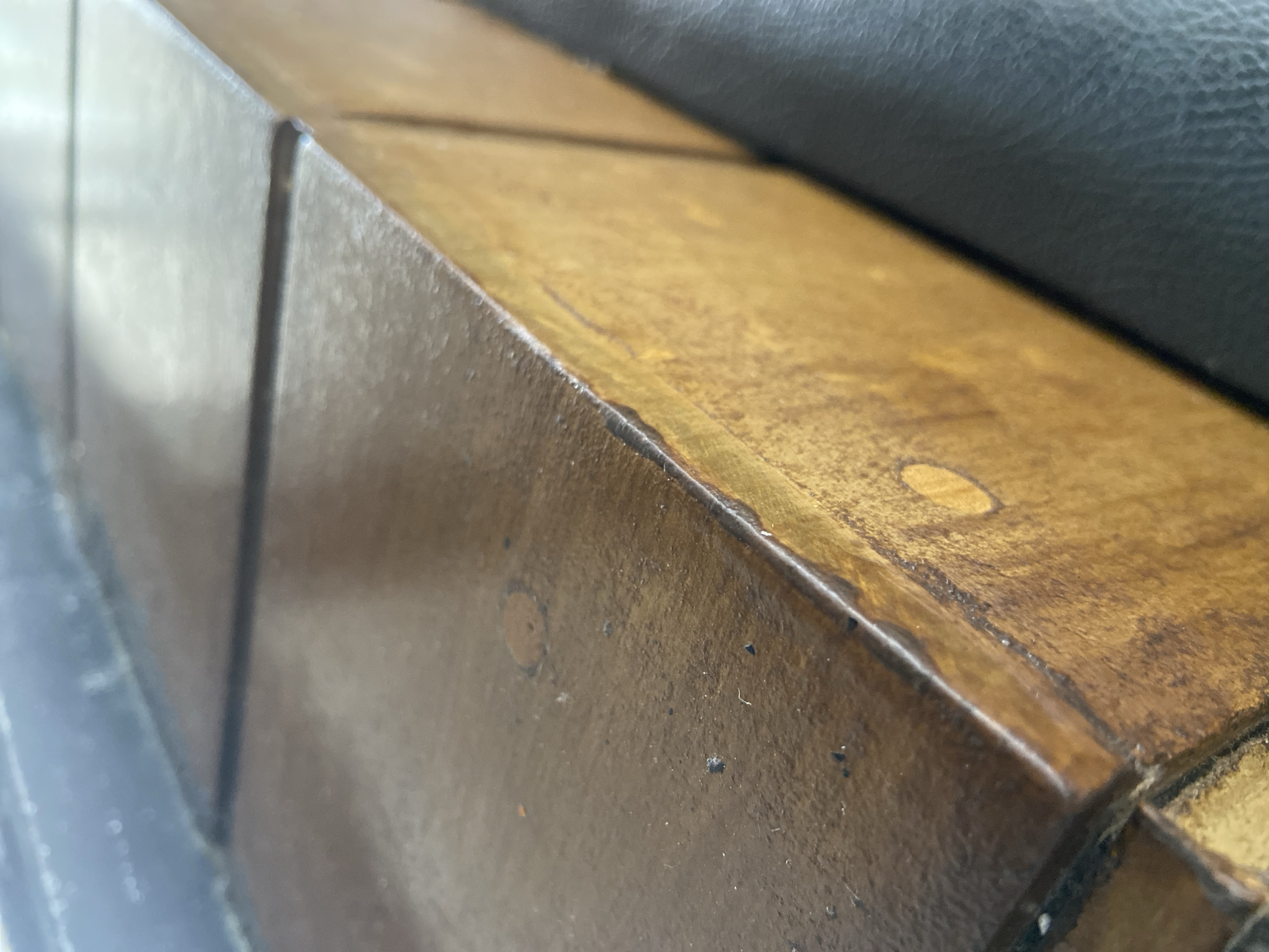 The image shows a close-up, side-angle view of a wooden object with a dark brown finish. The object appears to be a box or a segmented surface, with visible seams running horizontally and vertically across it. The surface has a glossy sheen, reflecting light from above, and shows some imperfections such as small dark specks and lighter circular marks, possibly knots in the wood. The edge of the object, where the top surface meets the side, is slightly raised and shows a lighter golden-brown color underneath the dark finish. To the right of the wooden object, a dark, textured surface, likely leather, is visible. The background on the left is out of focus and appears to be a dark, possibly reflective surface, with some indistinct white specks. There are no people or animals visible in the image, nor any context to suggest a specific location within the city of Bago, Indonesia. The lighting suggests an indoor environment with overhead light sources.