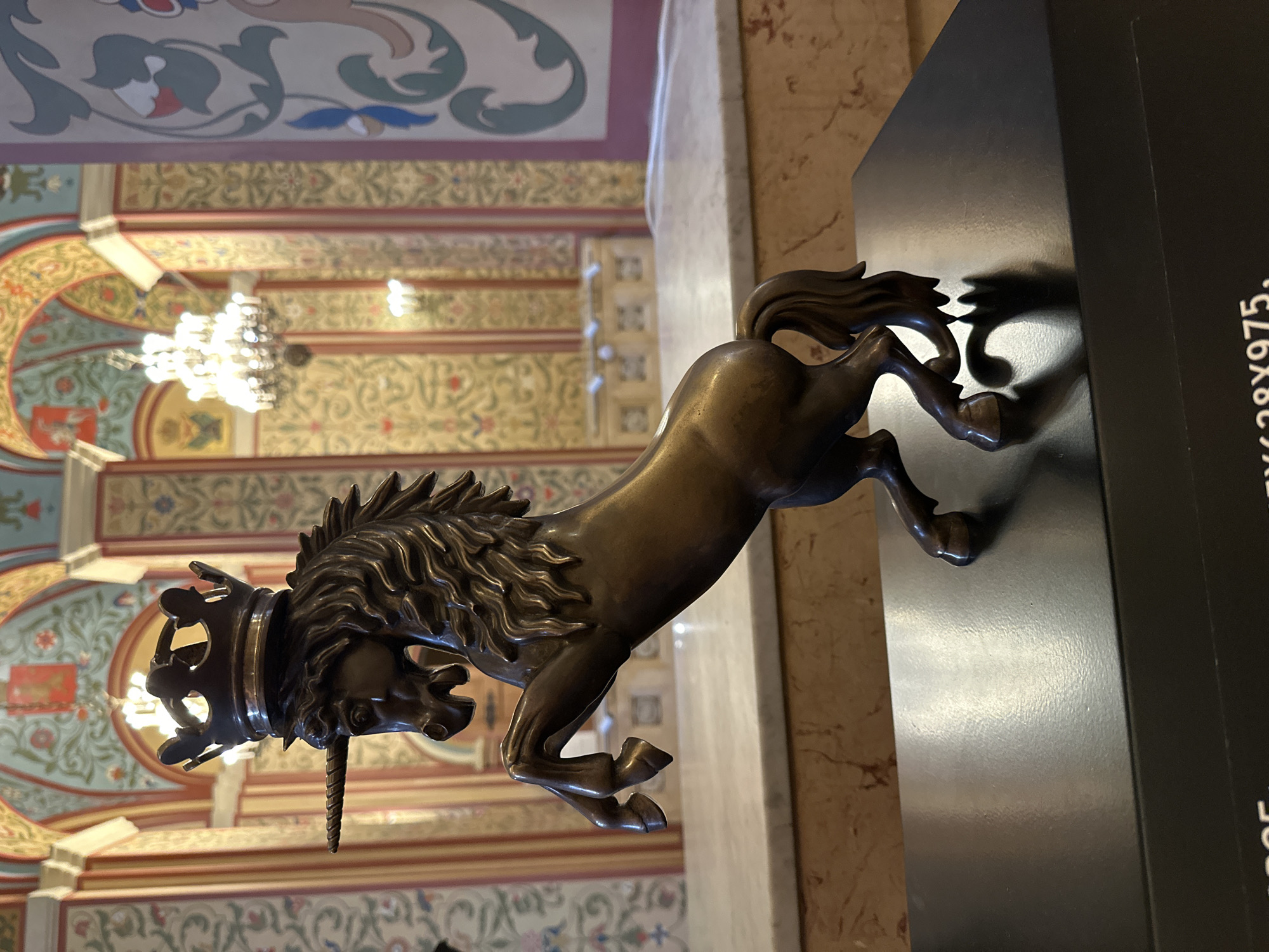 The image depicts a bronze sculpture of a unicorn wearing a crown, displayed indoors on a black pedestal. The unicorn is rearing on its hind legs, with its head turned slightly to the left. Its mane is flowing, and its horn is pointed upwards. The sculpture has a dark, metallic sheen.
The background suggests an opulent interior, possibly a museum or a grand hall. Arched ceilings with intricate paintings and decorative patterns are visible, along with a large, ornate chandelier. The walls are adorned with colorful murals and intricate wallpaper. Marble surfaces and polished wood accents contribute to the luxurious atmosphere.
The overall impression is one of historical significance and artistic craftsmanship, presented within a richly decorated setting. The style of the architecture and decor strongly suggests the interior of the State Kremlin Palace in Moscow, Russia, known for its grand halls and historical significance.