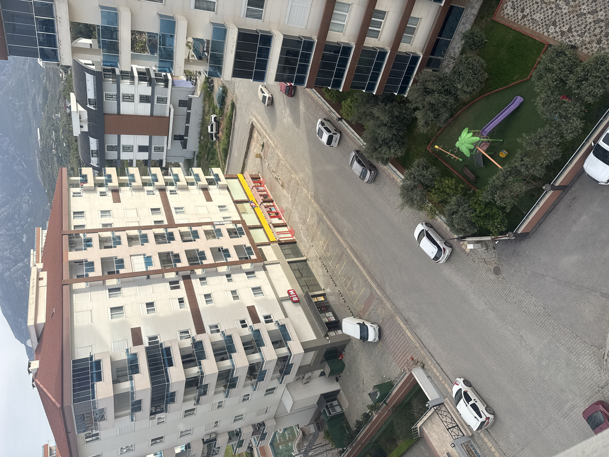 The image depicts an elevated perspective of a residential area in an unknown city. Several multi-story apartment buildings with white facades and glass balconies dominate the scene. A paved street runs between the buildings, populated by parked or moving cars, mostly white. On one side of the street is a small playground with green grass, trees, and play equipment including a purple slide. The red BIM logo is visible on the side of one of the buildings, indicating a supermarket. Distant mountains are visible in the background, partially obscured by the buildings.