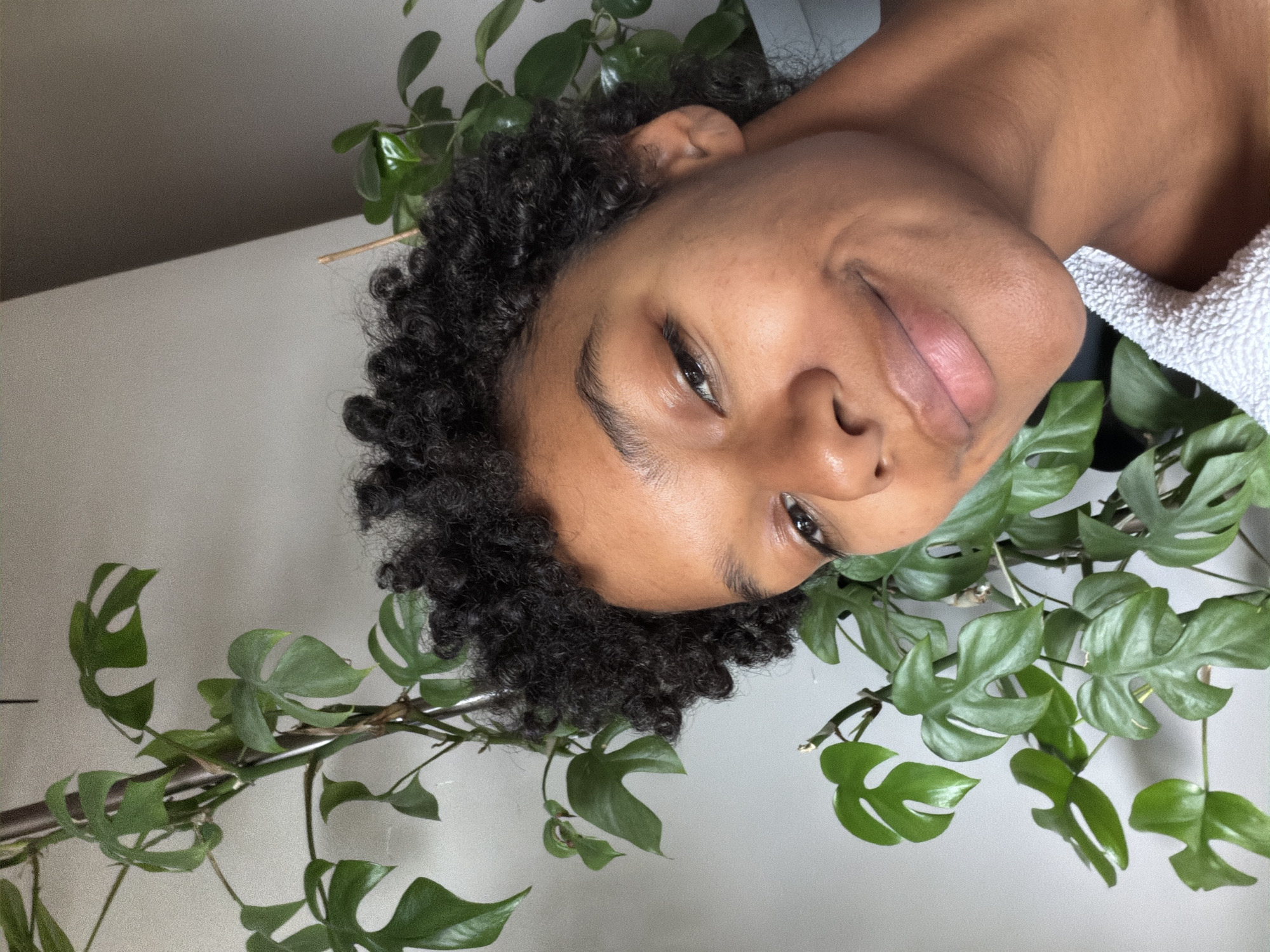The image displays a headshot of a young adult, with the frame oriented ninety degrees to the right. The person has dark, curly hair and brown skin, with a gentle smile and a direct gaze towards the camera. Their dark eyes are prominent, and their lips are slightly parted.
The person's head and shoulders are surrounded by lush green plants, specifically identifiable as a type of Monstera or Rhaphidophora, characterized by their unique fenestrated (split or holed) leaves. These plants are abundant, framing the person's face, hair, and neck, with stems and leaves visible on both sides and extending across the bottom and top (as per the image's rotated orientation). The foliage appears vibrant and healthy, suggesting an indoor environment where the plants are thriving.
A white, textured towel or similar fabric is draped over the person's right shoulder, providing a soft contrast to their skin and the green leaves. The background consists of a plain, light-colored wall, possibly off-white or light gray, with a subtle shadow line visible in the upper left corner, indicating an interior space. The lighting is soft and diffused, creating an even illumination without harsh shadows, which contributes to a calm and natural aesthetic. No text is visible in the image. The overall impression is one of natural beauty and tranquility, with the person interacting serenely with their plant surroundings in an indoor setting in Champs-sur-Marne, France.