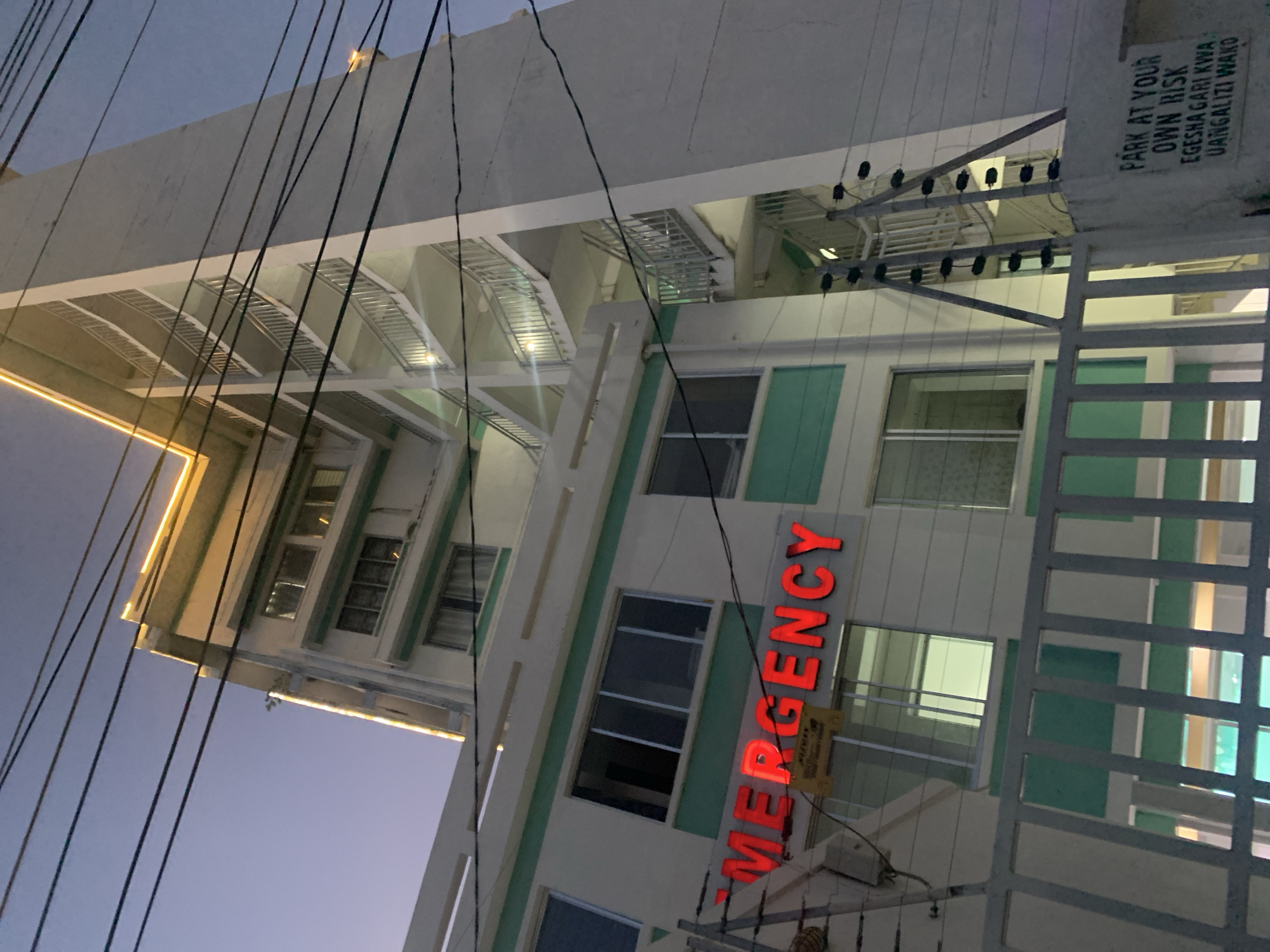 The image depicts a hospital building in Dar es Salaam, Tanzania. The word "EMERGENCY" is vertically displayed in red illuminated letters. The building is painted white with green accents around the windows. There is a sign with the text "PARK AT YOUR OWN RISK EGESHA GARI KWA UANGALIZI WAKO." Power lines are visible in the foreground. The sky is darkening, suggesting it is either dusk or dawn.