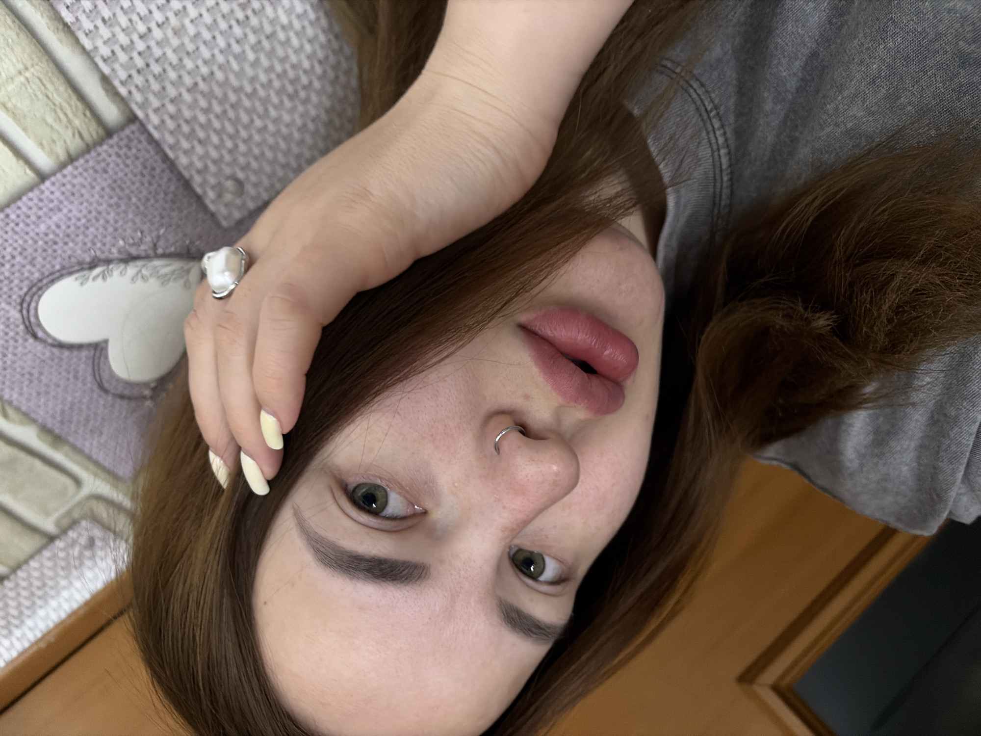 An overhead, slightly angled shot captures a young person lying down, looking directly at the camera. The subject has fair skin with visible freckles, brown hair, and striking green eyes. Their head is tilted, giving the image a unique orientation.
The person's left hand is raised towards their head, with manicured fingernails painted a light yellow shade. On their left ring finger, a silver ring adorned with a distinctive white, irregularly shaped pearl is visible. A silver septum ring pierces their nose. Their lips are full, with a natural pinkish hue, and slightly parted.
They appear to be wearing a grey t-shirt, which is partially visible on the right side of the frame. The background consists of a textured, patterned fabric, possibly a blanket or bedding, with a subtle heart-shaped outline design in a lighter shade. Below this, a wooden surface, possibly flooring or furniture, is visible. The lighting is soft and even, suggesting an indoor setting without strong directional light. There is no visible text in the image. The overall mood is relaxed and natural.