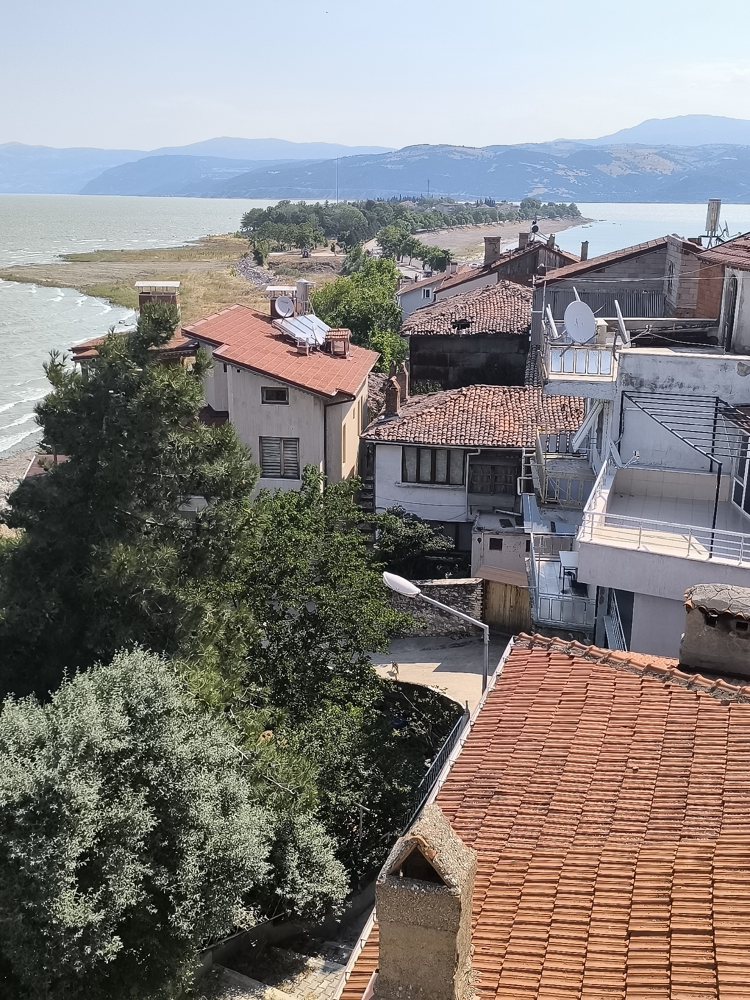 The image captures a scenic view of the city of Eğirdir, Turkey, overlooking the Eğirdir Lake. In the foreground, the terracotta tiled roofs of several houses are visible, with one prominent chimney made of weathered stone. Lush green trees partially obscure some of the buildings. Several houses feature solar panels on their roofs, suggesting a reliance on renewable energy.
Beyond the houses, the expansive Eğirdir Lake stretches towards the horizon, its waters a calm, pale blue under a clear sky. The shoreline is lined with a narrow strip of dry vegetation and scattered trees, indicating a warm, dry climate. In the distance, rolling hills and mountains form a gentle, undulating backdrop against the hazy sky. The overall impression is one of a peaceful, sun-drenched lakeside town.