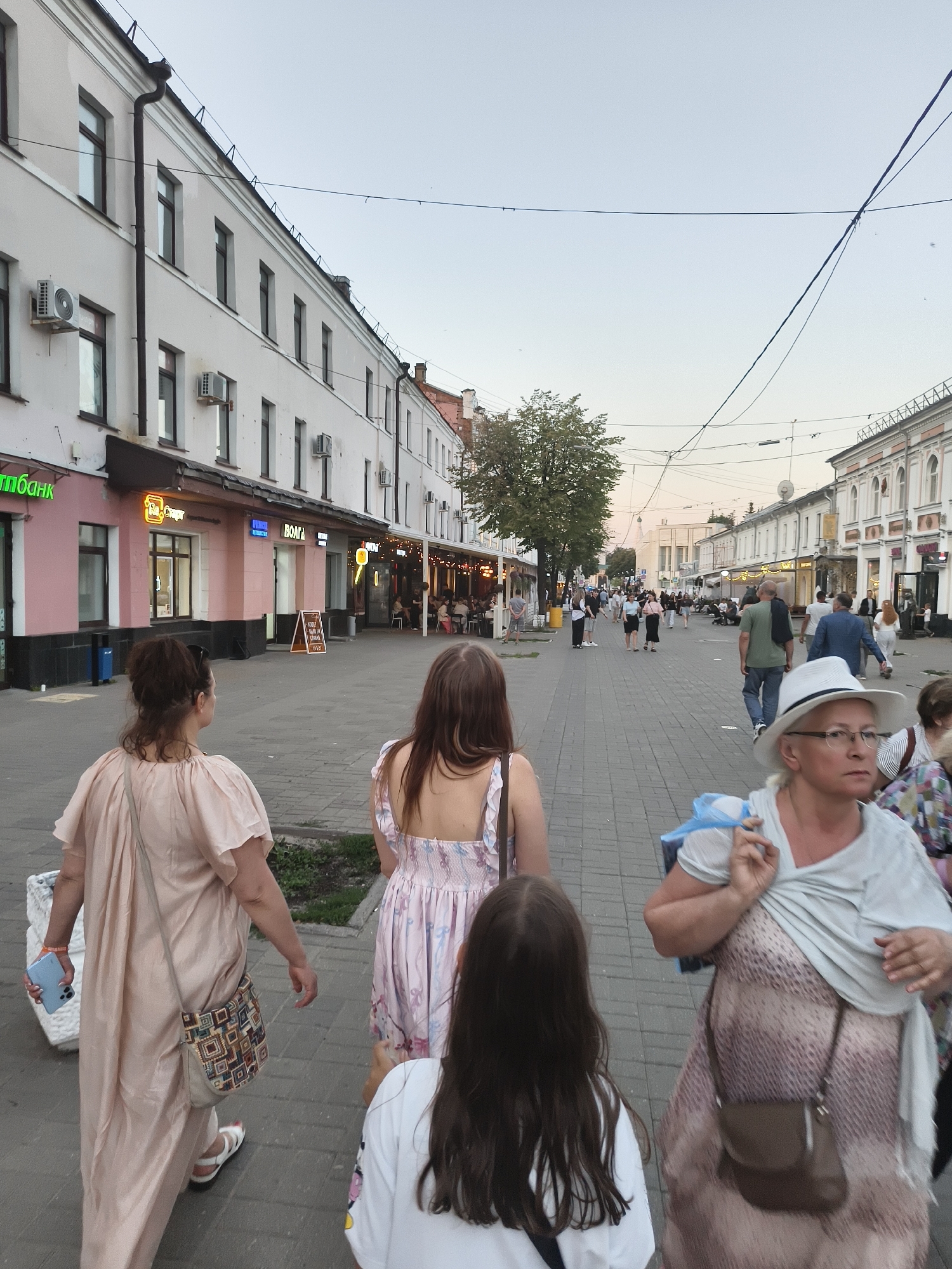 The image captures a street scene in Yaroslavl, Russia, likely in the evening as suggested by the lighting. The perspective is from behind a group of people walking away from the camera, looking down a pedestrian street lined with buildings.
In the foreground, three individuals are visible. Closest to the camera, from left to right, is a woman with her back to the viewer, wearing a light pink, flowing dress and carrying a light blue smartphone. Next is a young girl with long dark hair, also facing away. Further to the right, a woman in a patterned dress and a white hat is walking towards the camera, holding a shopping bag. Behind her, other pedestrians are visible, including a man in a green t-shirt and a man in a blue suit.
The street itself is paved with grey interlocking bricks. On either side of the street are multi-story buildings with storefronts at ground level and windows on upper floors. Some of the storefronts have illuminated signage. The buildings on the left appear to be primarily pink and white, with air conditioning units visible on the facades. On the right, the buildings are predominantly white with decorative architectural elements.
Trees are present along the street, and in the distance, the street appears to continue, with more buildings and lights suggesting a commercial area. Electrical wires are strung across the sky, typical of an urban environment. The sky is a pale blue, transitioning to a lighter hue near the horizon, indicating either early morning or late evening. People are seen strolling, sitting at outdoor cafe tables, and generally engaging in leisurely activity. The overall atmosphere is that of a busy, pleasant urban pedestrian zone.