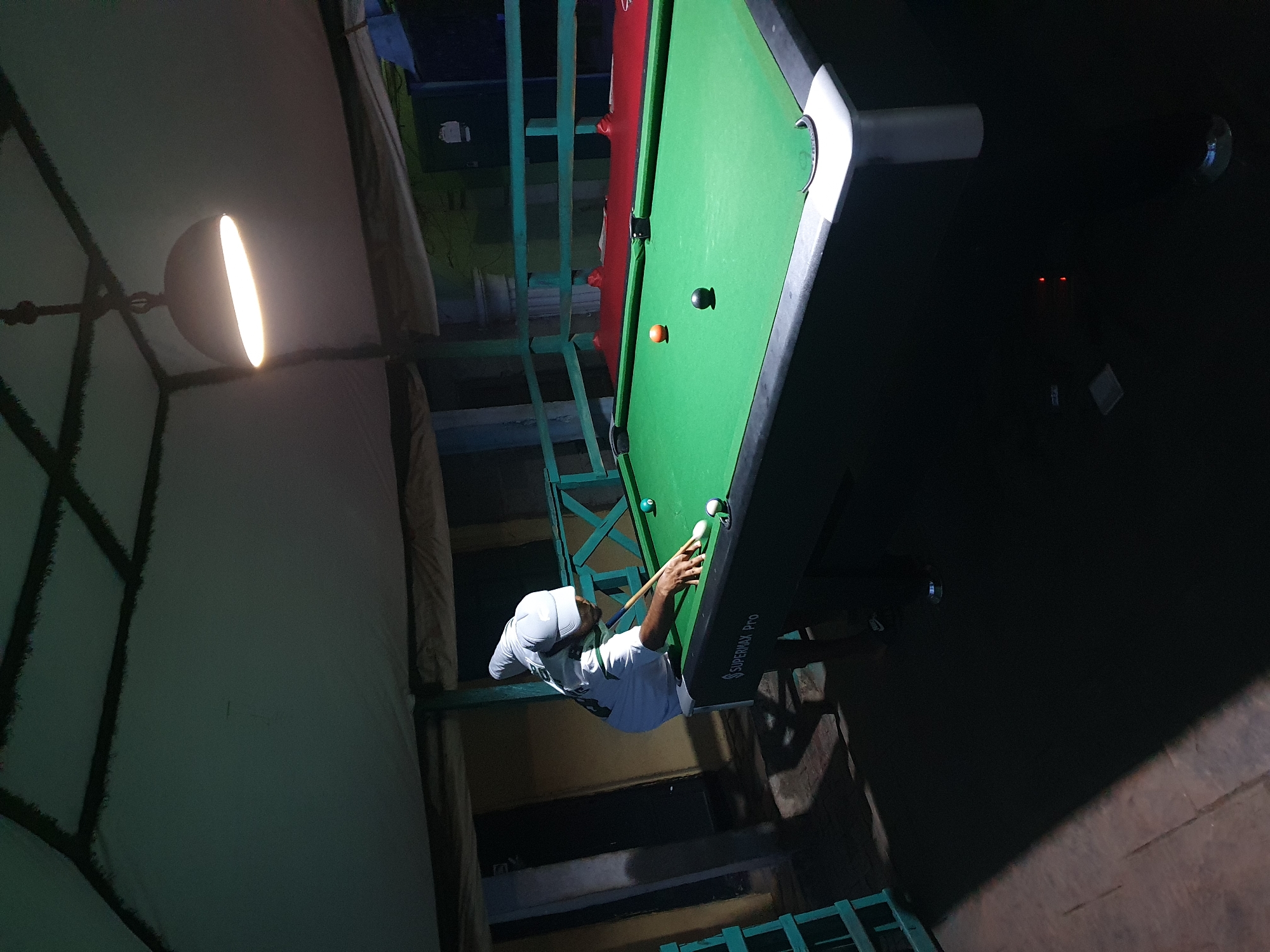 The image shows a man playing pool indoors under artificial light. He is wearing a white t-shirt and a white hat, and is positioned at the end of the pool table. There are several pool balls on the green table, along with a cue stick in his hands. The table is labeled "SUPERMAX PRO". A light fixture hangs from the ceiling, illuminating the scene. The surrounding environment appears to be a room with textured walls and green metal framework. The specific location in Lokoja, Nigeria cannot be determined from the image.