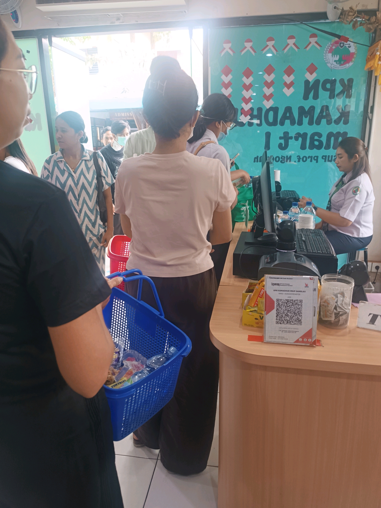 The image depicts a queue of people inside a bright establishment in Denpasar, Indonesia. The queue appears to be for a payment or service counter. In the foreground, a person wearing a black top is holding a blue plastic basket filled with items, suggesting they are a customer. Behind them, another person in a light-colored top is visible. Further back, several other individuals are waiting, including one wearing a mask and a patterned shirt, and another in a light blue uniform.
The background features a large turquoise-colored board with text and graphics, likely advertising or informational. A computer monitor, keyboard, and a QR code scanner are prominent on the counter where a person in a light uniform is seated, presumably a cashier or service agent. Water bottles and a container with cash are also on the counter. The overall environment suggests a retail or administrative setting. The lighting indicates it is daytime, with natural light entering from an open doorway or window on the left side of the image.