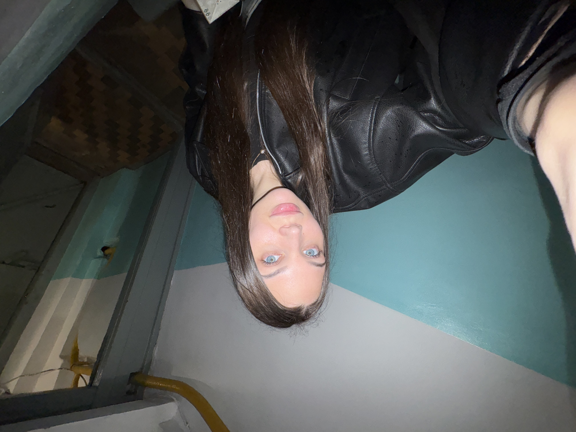 A selfie photograph taken in a stairwell in the city of Chaikovsky, Russia shows a young woman with light skin, brown hair, and blue eyes. She is wearing a black leather jacket. The stairwell has light blue and white painted walls. A yellow electrical cord is visible in the lower left of the image. The image is rotated 180 degrees.