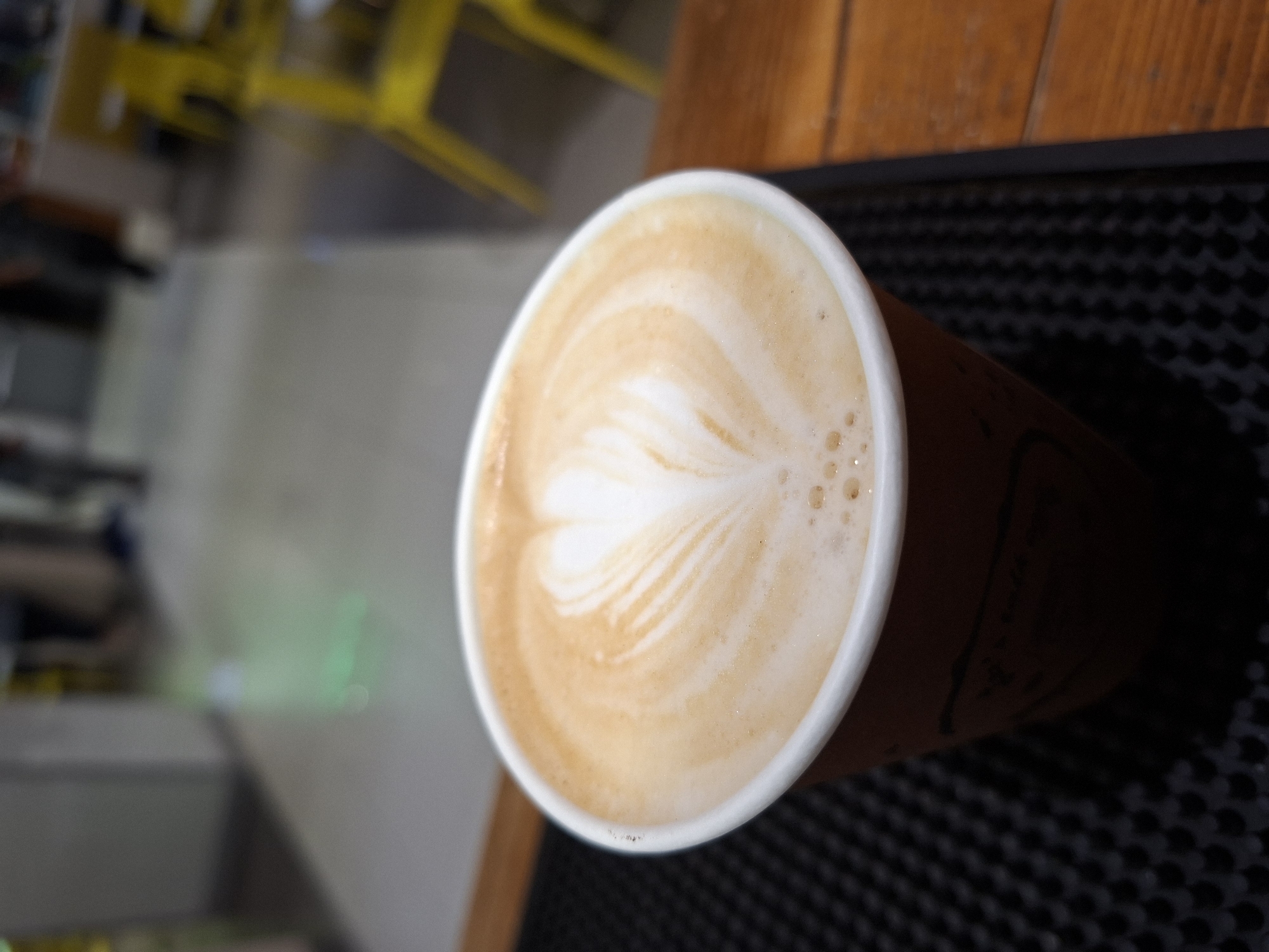 A high-angle shot shows a latte in a disposable cup with latte art. The latte is placed on a black, textured mat, possibly a bar surface. A blurred background suggests an interior setting, with hints of yellow chairs and neutral wall colors. The cup has a brown exterior. The latte art displays a heart-shaped design formed with foam. The location is unspecified in the city of Plovdiv, Bulgaria, and no people are depicted.