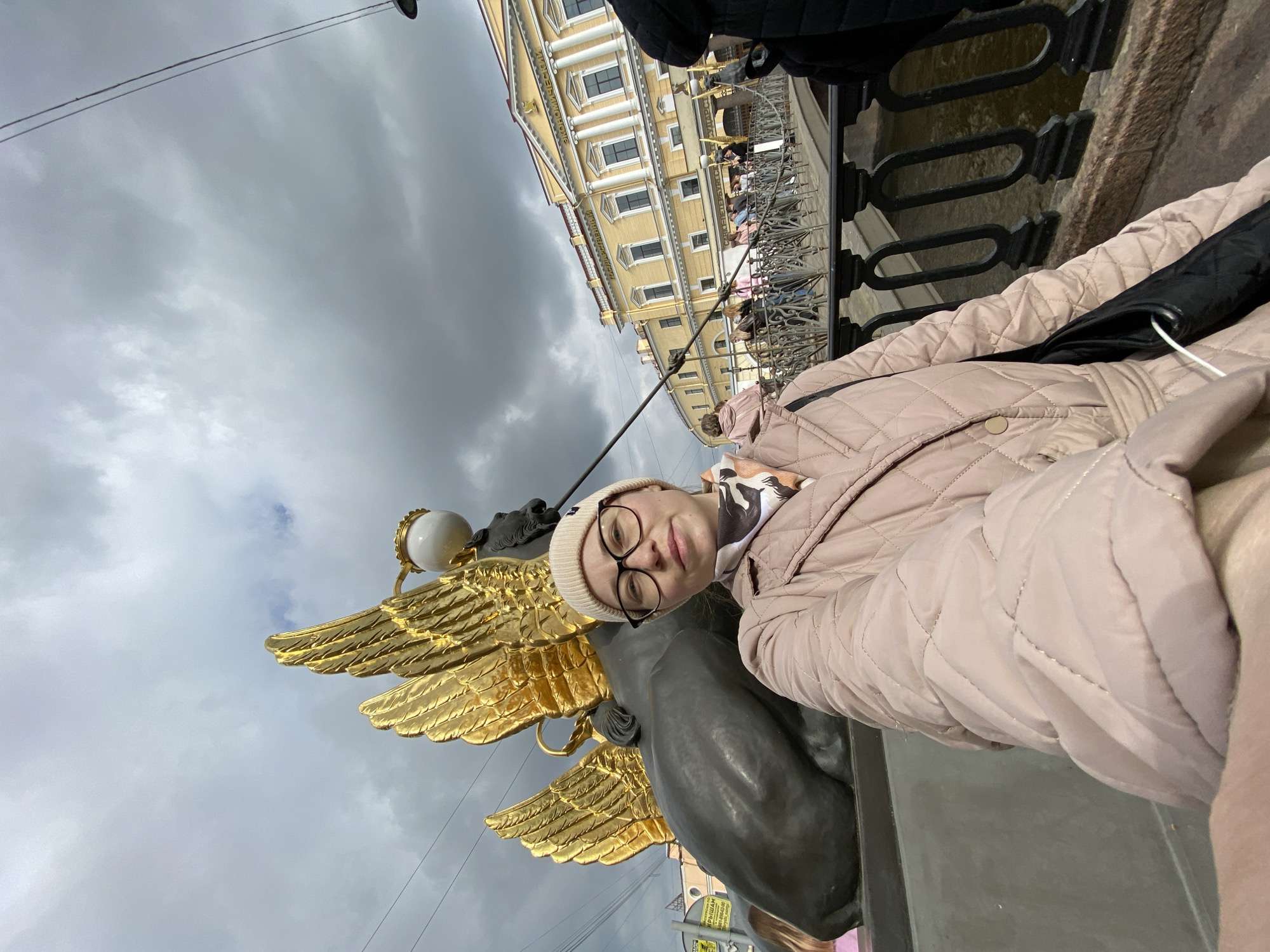 A woman wearing a beige jacket, beanie, and glasses poses next to a griffin statue on the Bank Bridge in Saint Petersburg, Russia. The gold-winged griffin stands on a plinth against a cloudy sky. In the background is the yellow façade of a building across the canal, with a black metal bridge railing separating the viewer from the water. Other people are visible on the bridge behind the railing.