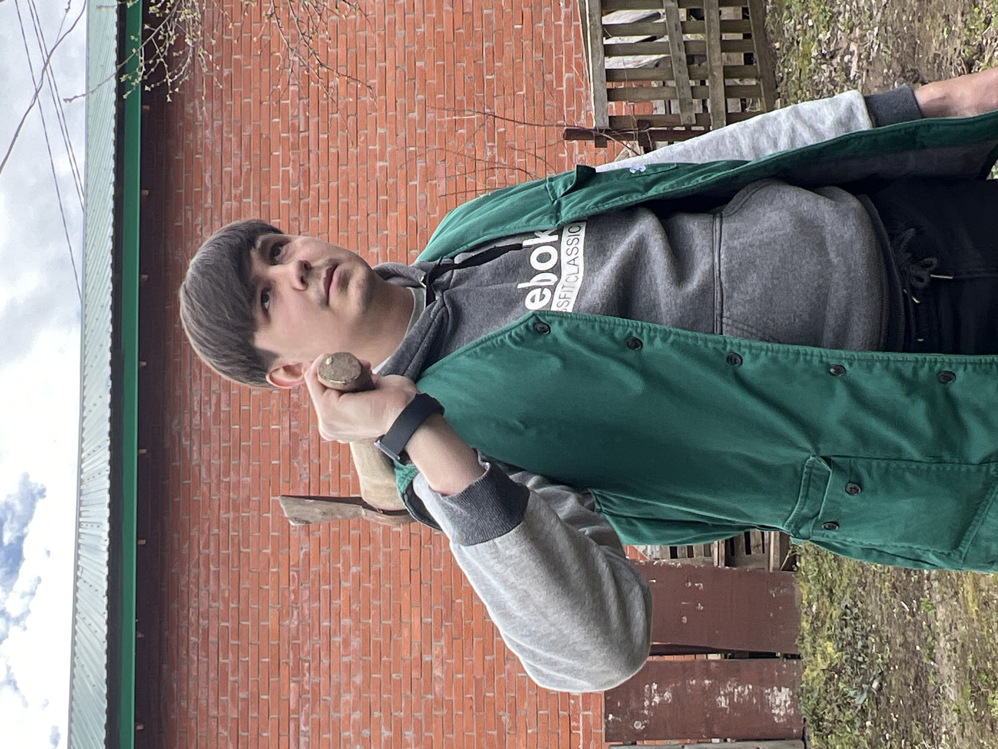 A young man stands outdoors in Chaikovsky, Russia, holding a small axe. He is wearing a grey hoodie with the brand "Reebok" printed on it, a green jacket, black pants, and a black smartwatch on his left wrist. He is looking upwards and slightly to his left. The backdrop consists of a red brick wall on the left side of the frame and a wooden fence on the right. The sky is visible at the top left, suggesting a cloudy day. The surroundings appear to be a yard with some vegetation present.