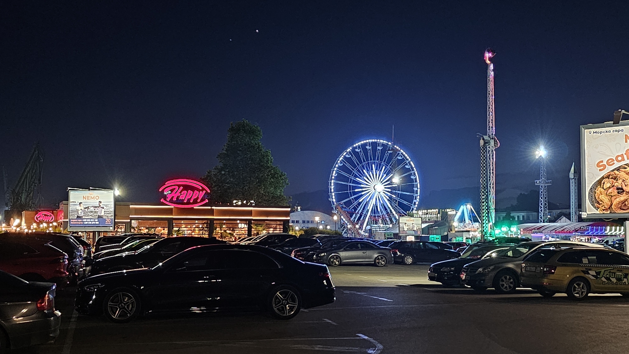 The image depicts a nighttime scene at the Varna Sea Garden's amusement park in Varna, Bulgaria. Dominating the skyline is a large, brightly lit Ferris wheel with blue lights, and a tall amusement ride with flashing lights. In the foreground, a parking lot is filled with various cars, including a yellow taxi. Behind the parking lot, a building with a neon pink "Happy" sign is visible, likely a restaurant. Further in the background, a large billboard advertising "Seafood" is illuminated. The sky is dark, with two faint celestial bodies visible. The overall impression is that of a lively evening at an entertainment venue.