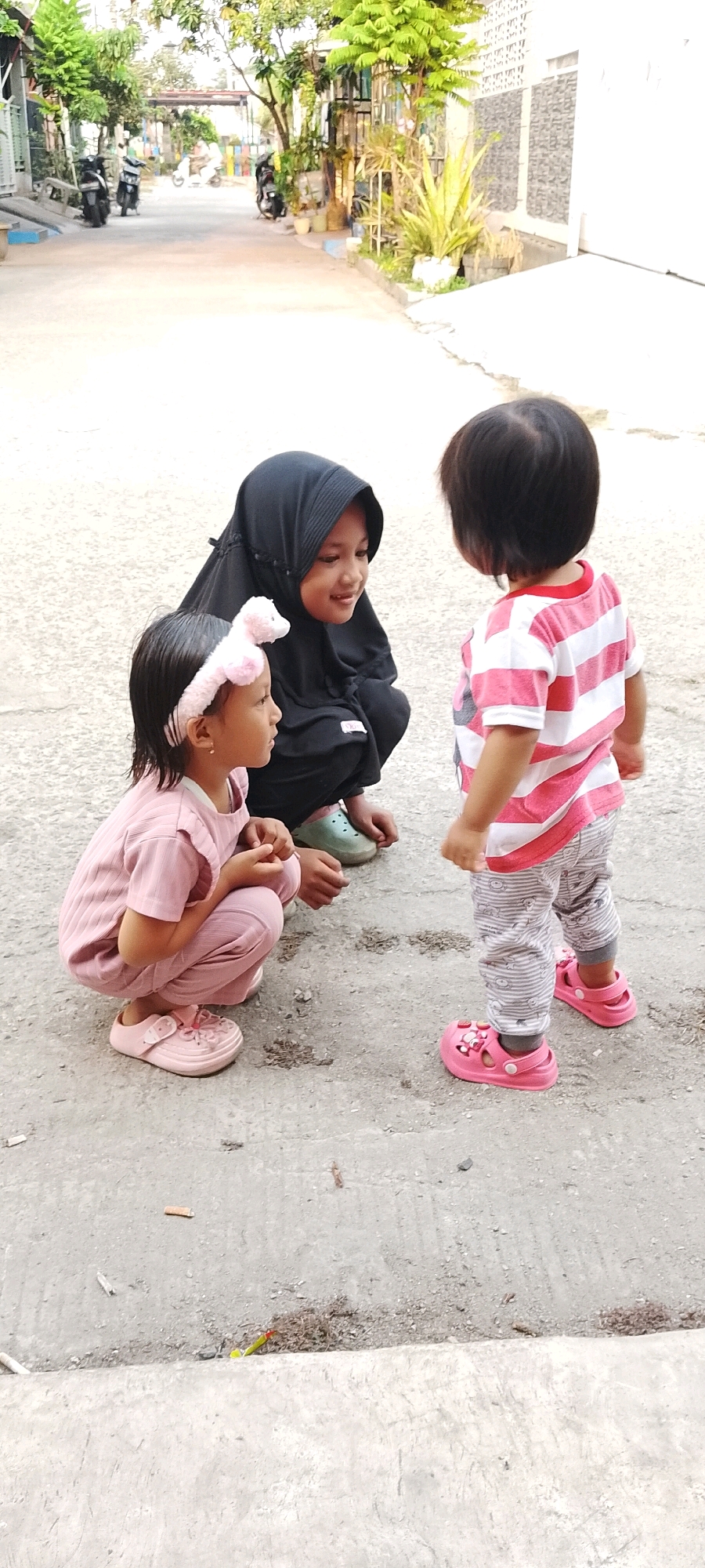 In the city of Satriamekar, Indonesia, three young children are interacting on a paved outdoor area. The child on the left, wearing a pink outfit and a pink headband with bunny ears, is crouched down, facing the child on the right. The child in the middle is wearing a black hijab and is also crouched, looking towards the child on the right with a smile. The child on the right, facing away from the camera, is wearing a pink and white striped t-shirt and patterned pants, and is standing. The ground is concrete with some gravel and debris. In the background, there is a street with parked motorcycles, trees, and buildings.