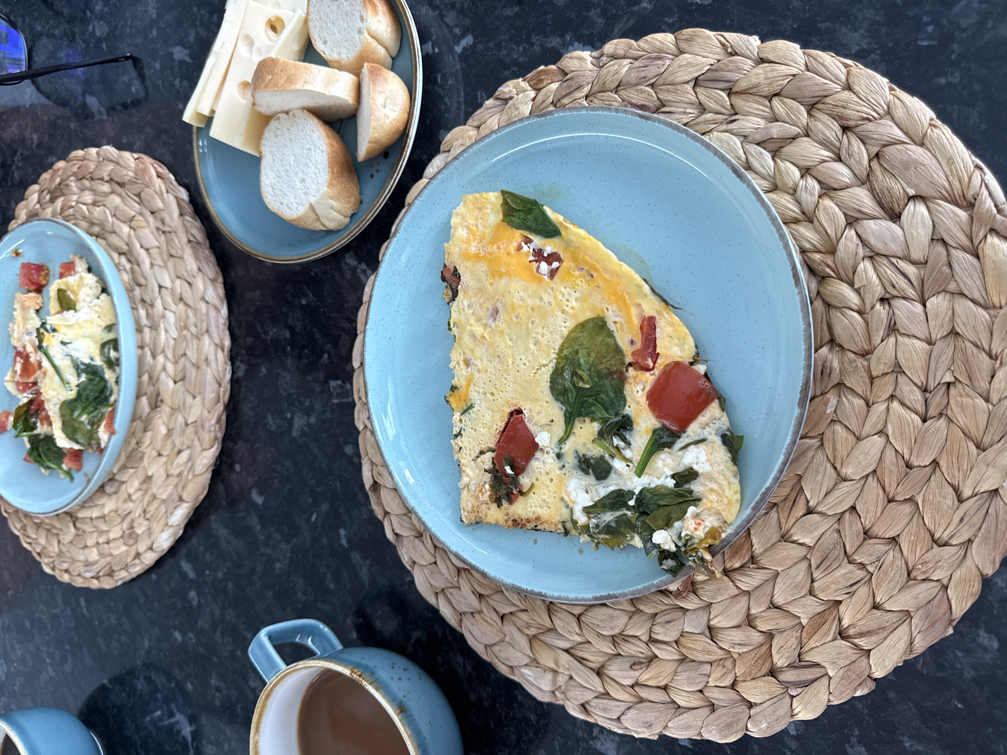 The image shows a breakfast scene on a dark countertop. A slice of frittata, containing spinach, tomatoes, and cheese, rests on a light blue plate, which sits on a round woven placemat. Nearby, a smaller blue plate holds slices of baguette and cheese. In the lower left corner, there is a blue mug filled with a brown liquid, possibly coffee or tea. A pair of glasses is near the plate with bread and cheese. Another similar plate of the frittata is on a woven placemat. The scene appears to be indoors. There is no indication of this being in Sochi, Russia.