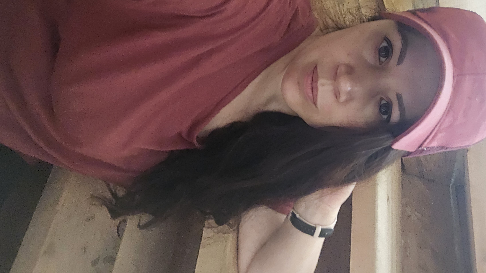 In this medium shot, a woman with dark hair, wearing a pink baseball cap and a maroon t-shirt, poses for a selfie. She is leaning against a wooden staircase, with her right arm bent and her hand resting on her head. Her gaze is directed at the camera, and she has a neutral expression. A black and white fitness tracker is visible on her left wrist. The background consists of the wooden steps of a staircase, suggesting an indoor setting, possibly a home. The lighting is natural, casting soft shadows. There is no indication of the location being in Ермолаево, Russia; the context is purely speculative.
