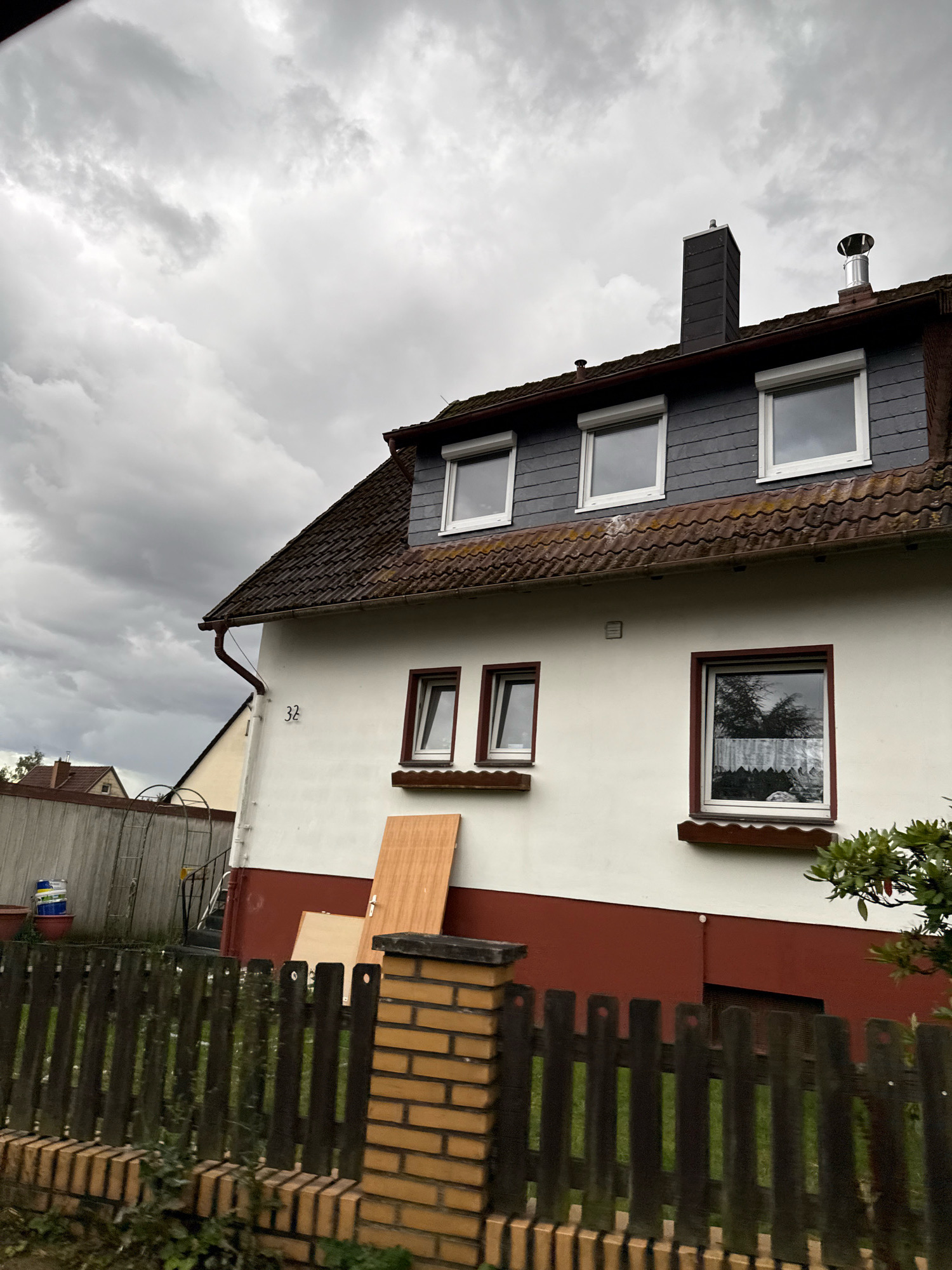 This is a medium-shot, eye-level photograph of a two-story house on a cloudy day in the town of Bad Sachsa, Germany. The house is white with a red-painted foundation and a dark grey, shingled roof. It has several windows, some with white shutters. A chimney with a silver cap is visible on the roof. In front of the house, there is a dark wooden fence with brick posts. Leaning against the side of the house are two wooden planks, possibly for construction or renovation. The sky is overcast with heavy grey clouds, suggesting impending rain. The house number "32" is visible on the white exterior wall. In the background, part of another house and a wooden fence can be seen.