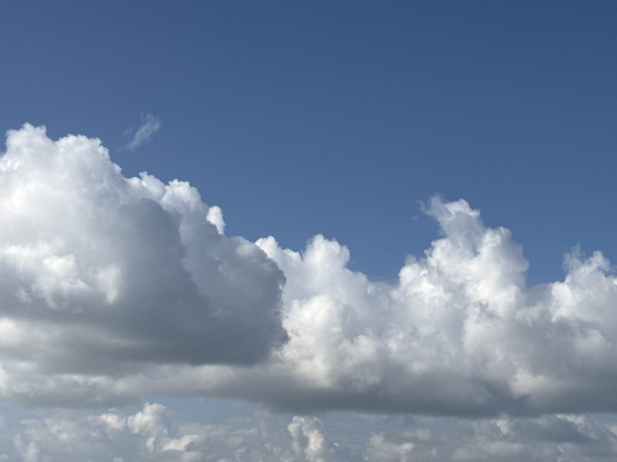 A wide-angle shot captures a bright blue sky filled with a variety of clouds. A large, dominant cloud formation stretches across the lower half of the image, with lighter, fluffy clouds scattered above and below. The sky appears clear and sunny, typical of an outdoor scene. The image is a photograph of the sky.